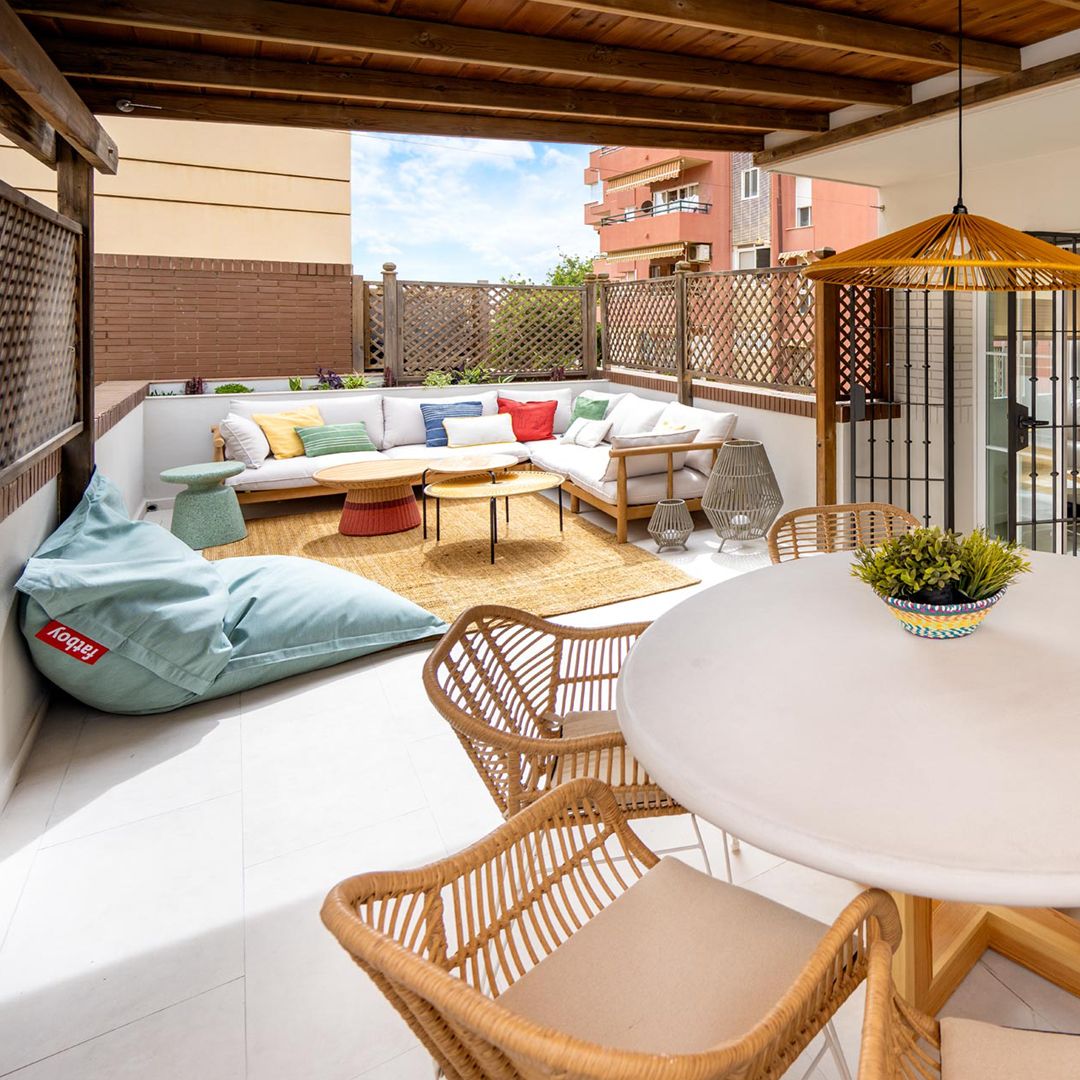 Patio al aire libre en la azotea amueblado con un sofá seccional blanco, coloridos cojines, dos pufs, una mesa de madera y una alfombra estampada de Vivienda Pedregalejo, rodeado por una valla de privacidad.