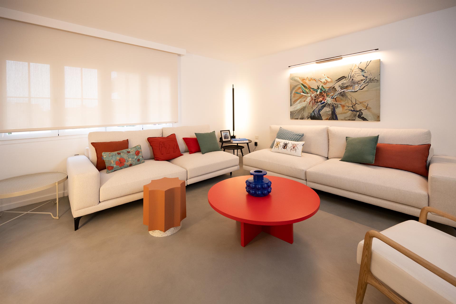 vivienda-pedregalejo-proyecto-interiorismo-viviendas-blancacano-016 Una sala de estar moderna con un gran sofá seccional blanco, cojines coloridos, una mesa de centro redonda roja y arte abstracto en la pared.