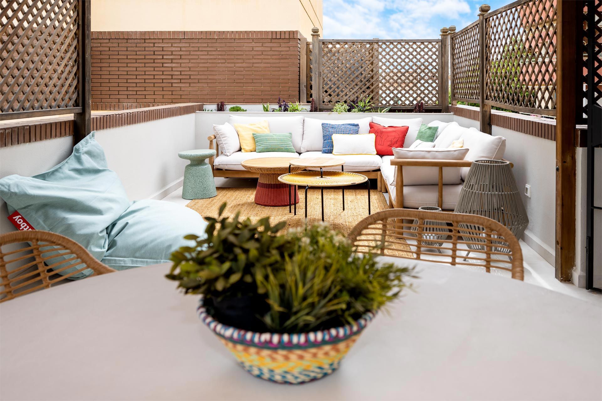 Acogedor patio en la azotea con muebles de mimbre, cojines coloridos y una mesa central de madera, rodeado por una celosía que protege la privacidad.