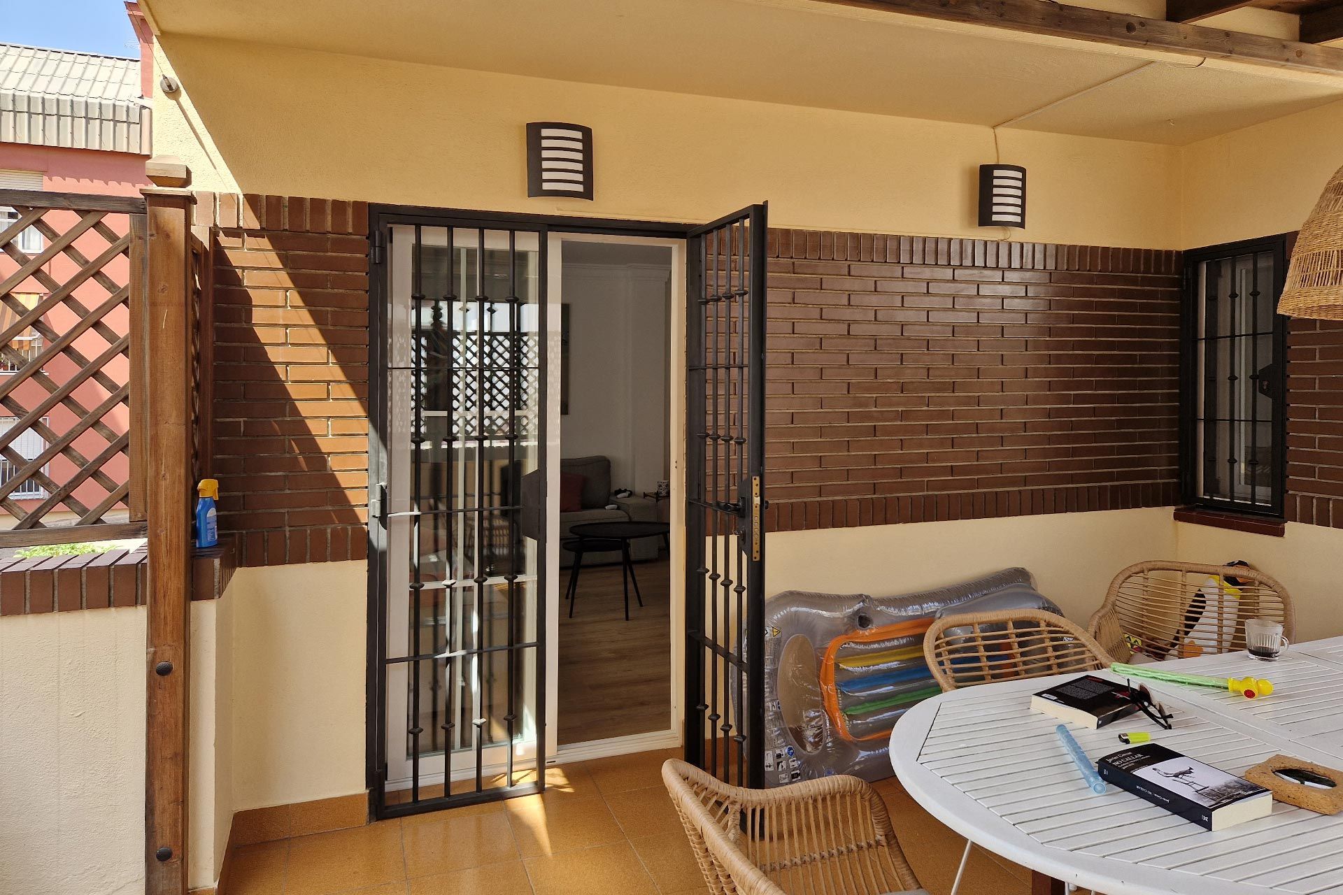 Un acogedor patio cubierto con una mesa blanca, dos sillas de mimbre, paredes de ladrillo y una puerta abierta que conduce al interior de una casa.
