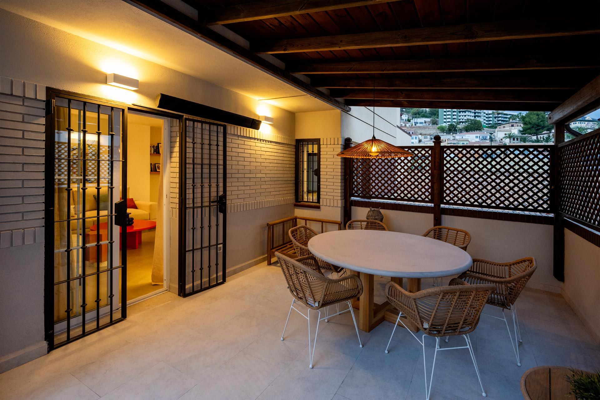 Una amplia terraza al atardecer con una mesa redonda con sillas, realzada por una iluminación cálida, junto a las puertas corredizas abiertas de una casa.