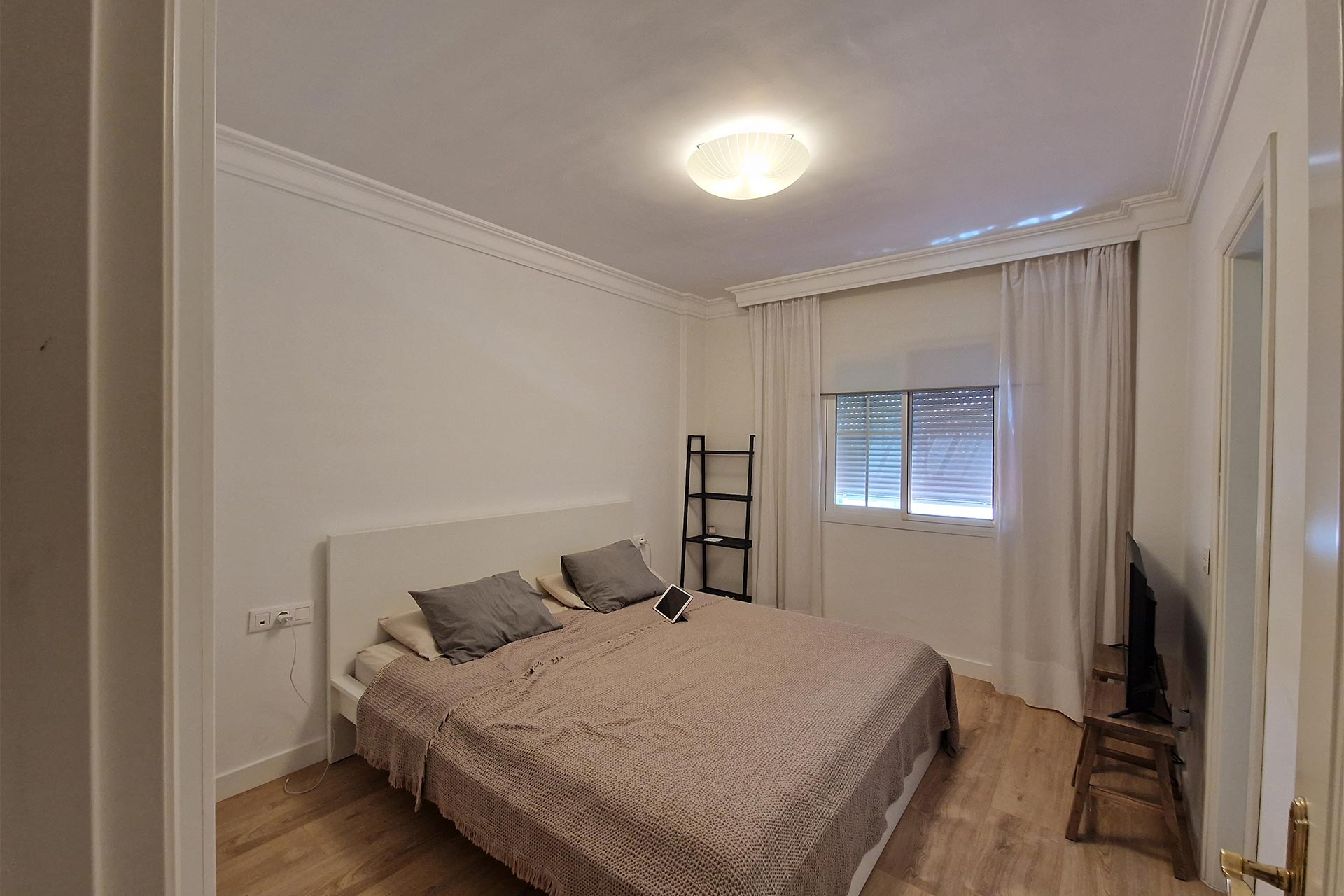 Un dormitorio minimalista con cama de matrimonio, ropa de cama gris, una pequeña estantería negra con forma de escalera, cortinas blancas y una lámpara de techo.