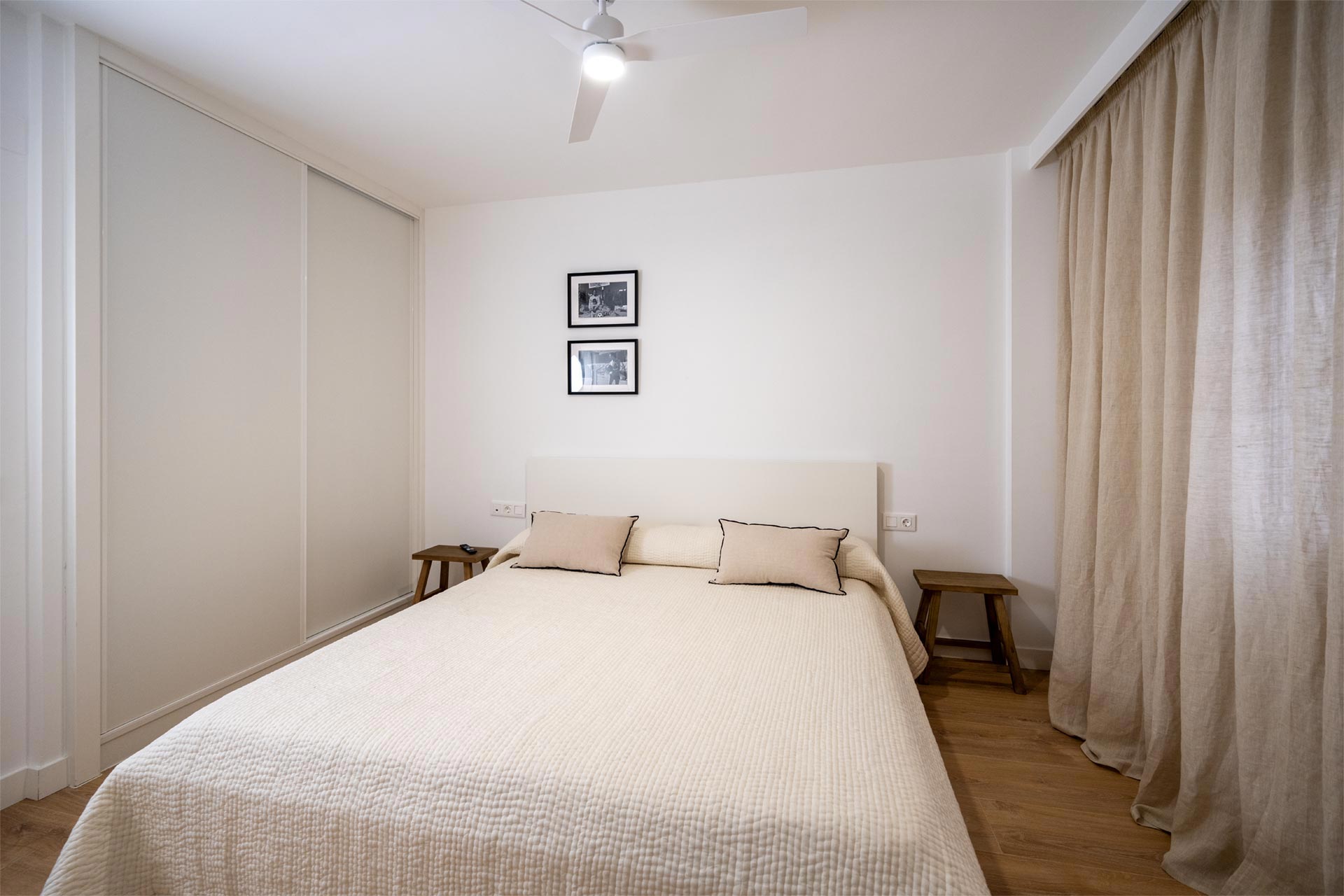 Un dormitorio moderno con una cama grande, ropa de cama beige y fotografías enmarcadas encima de la cabecera. hay cortinas a la derecha y un escritorio a la izquierda.