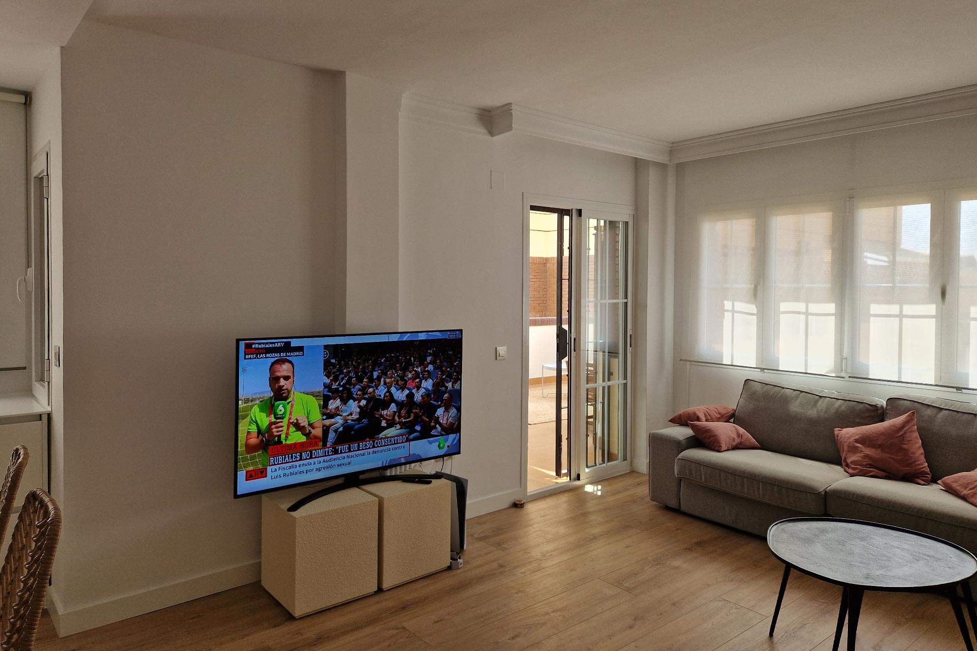 Sala de estar moderna con un sofá gris, televisión montada que muestra un canal de noticias y puertas abiertas del balcón que dejan entrar luz natural.