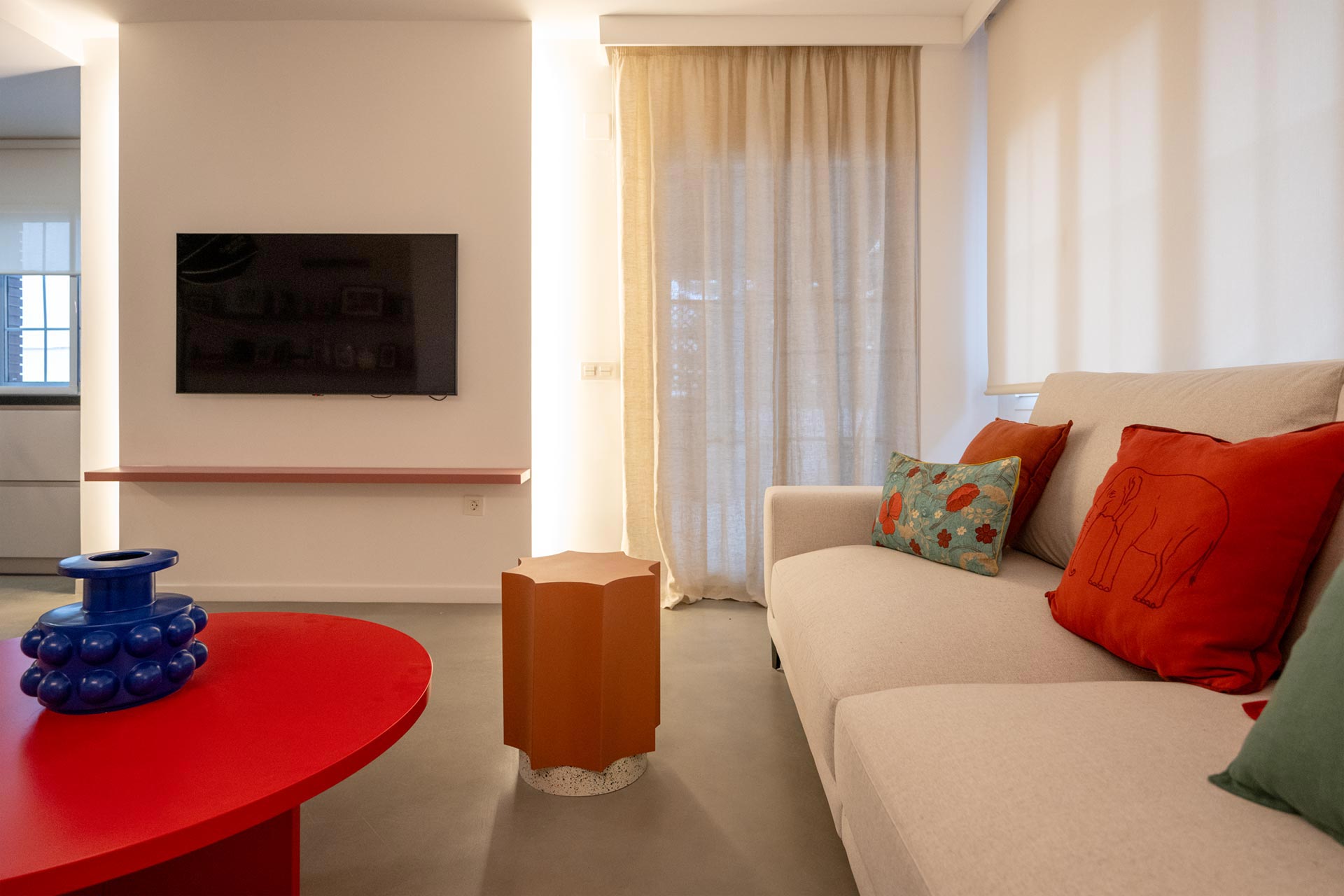 Una sala de estar moderna con un sofá blanco adornado con almohadas de colores, un televisor grande montado en la pared, una mesa de centro roja y un adorno azul único.