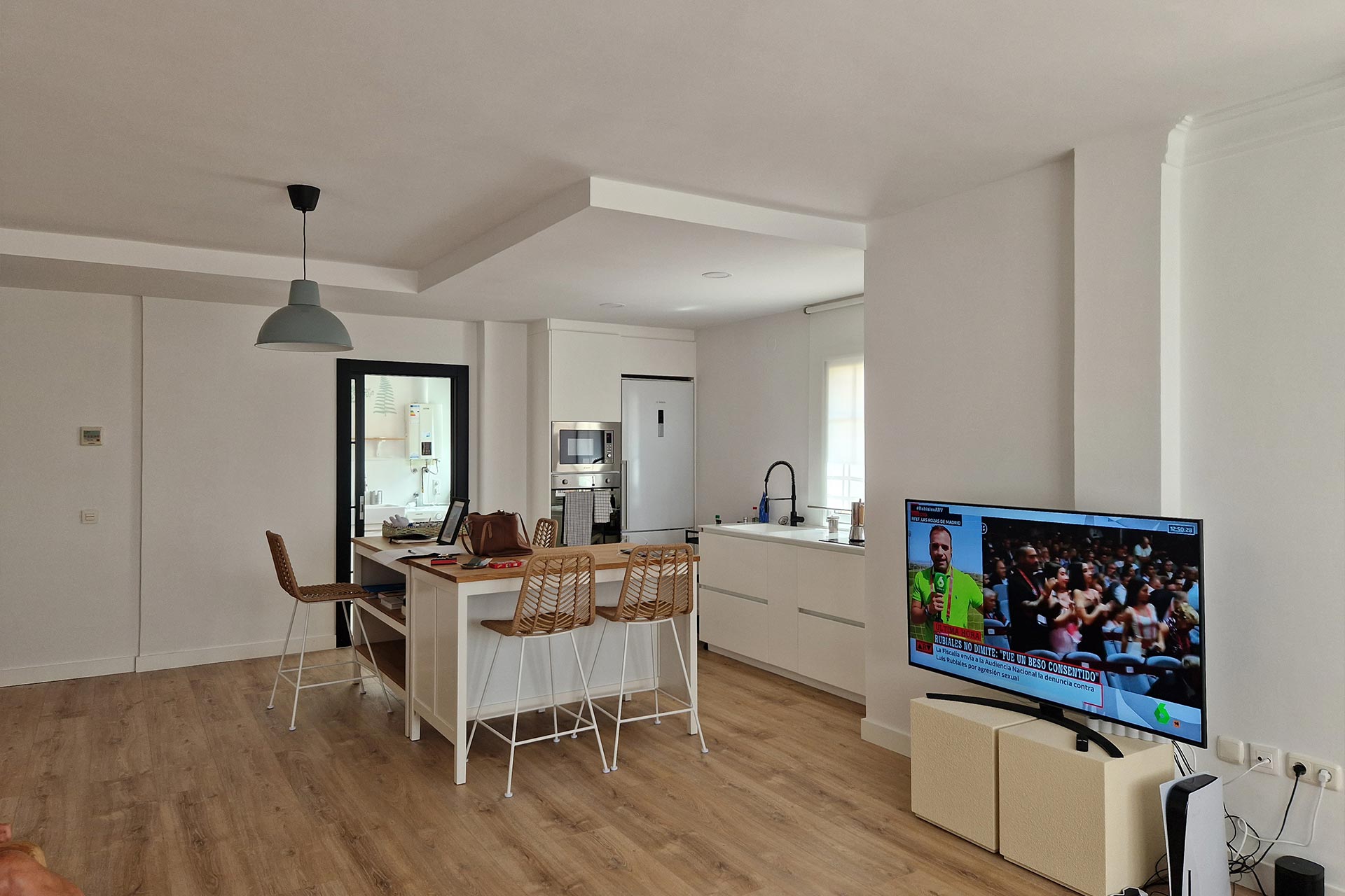 Cocina y comedor modernos con gabinetes blancos, una isla con taburetes, un escritorio con una computadora portátil y una sala de estar con un televisor que muestra un partido deportivo.