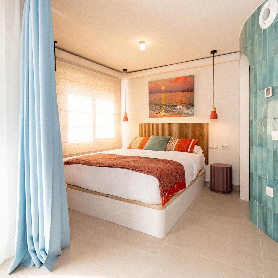 Un dormitorio con una cama grande, cortinas azules, luces colgantes de color naranja, una manta estampada, un cuadro colgado en la pared y una pared con detalles de azulejos.