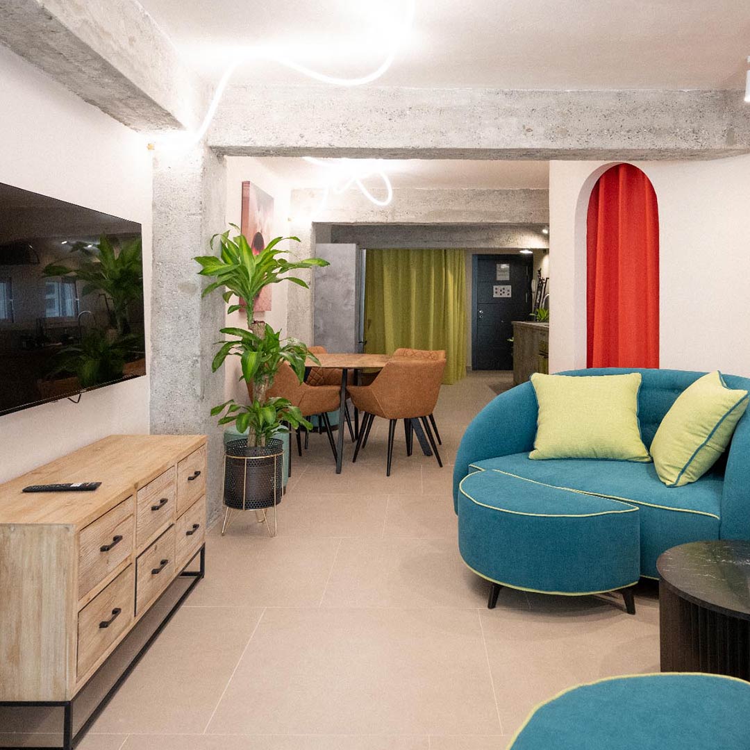 Una moderna sala de estar y comedor con un sofá redondo turquesa con cojines amarillos, una cómoda de madera, un televisor montado en la pared, plantas en macetas y una mesa de comedor con cuatro sillas marrones.