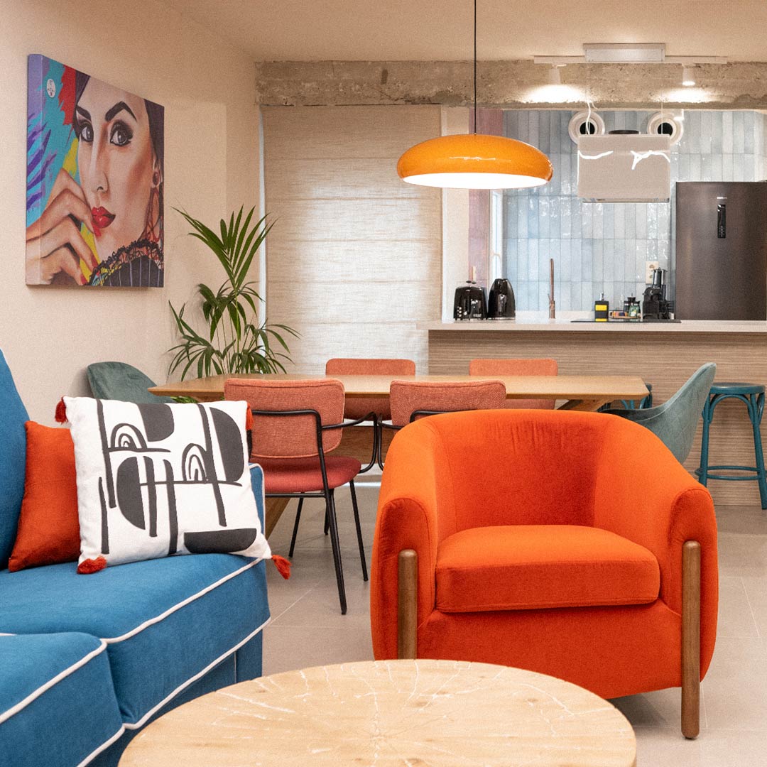 Una sala de estar moderna con un sofá azul con cojines estampados, un sillón naranja y una mesa de centro redonda de madera. El comedor tiene sillas naranjas y una lámpara colgante, con una cocina al fondo.