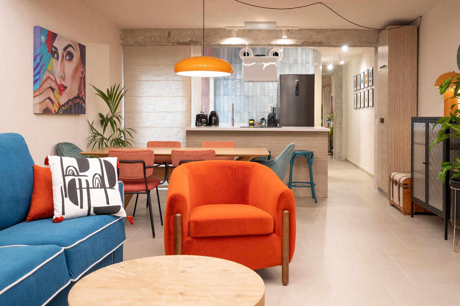 vivienda-calle-peña-proyecto-interiorismo-viviendas-turisticas-blancacano-d-19 Una sala de estar moderna y bien iluminada con muebles coloridos, que incluyen un sillón naranja y un sofá azul. Una mesa de comedor con sillas y una cocina con barra se encuentran al fondo.