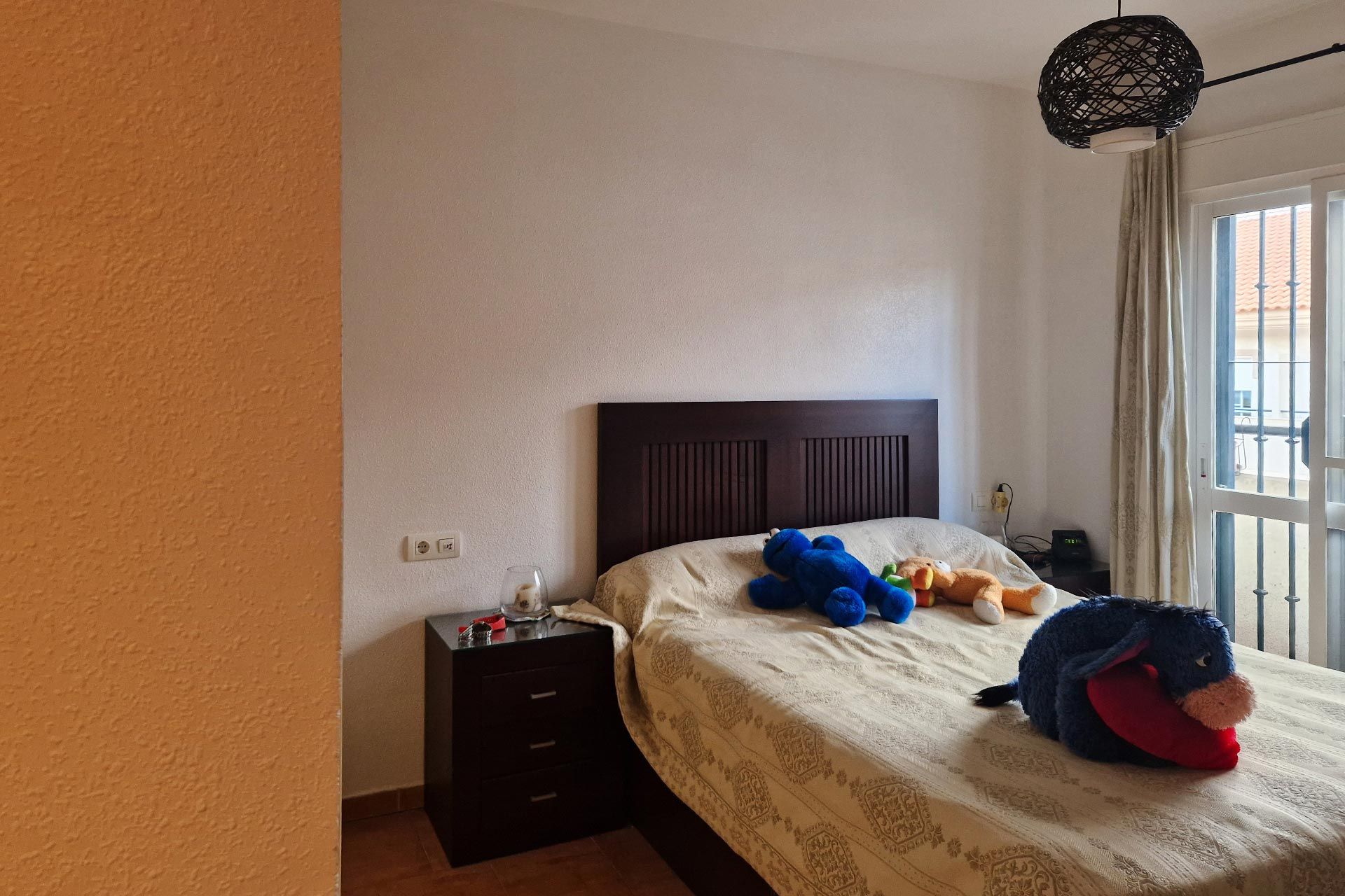Un dormitorio con una cama de madera, peluches sobre la cama, una mesita de noche con objetos, una ventana con cortinas y una lámpara colgando del techo.