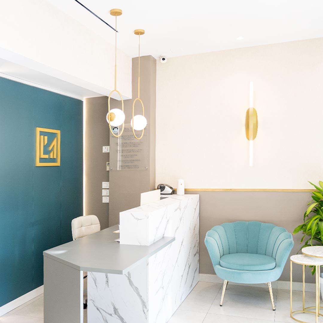 reforma-clinica-dental-blancacano-00 La recepción de Clínica Muñoz cuenta con un mostrador de mármol, una silla azul y luminarias modernas. La pared está pintada en tonos verde azulado y beige con un logotipo con detalles dorados. Una planta en maceta añade vegetación a este elegante espacio.
