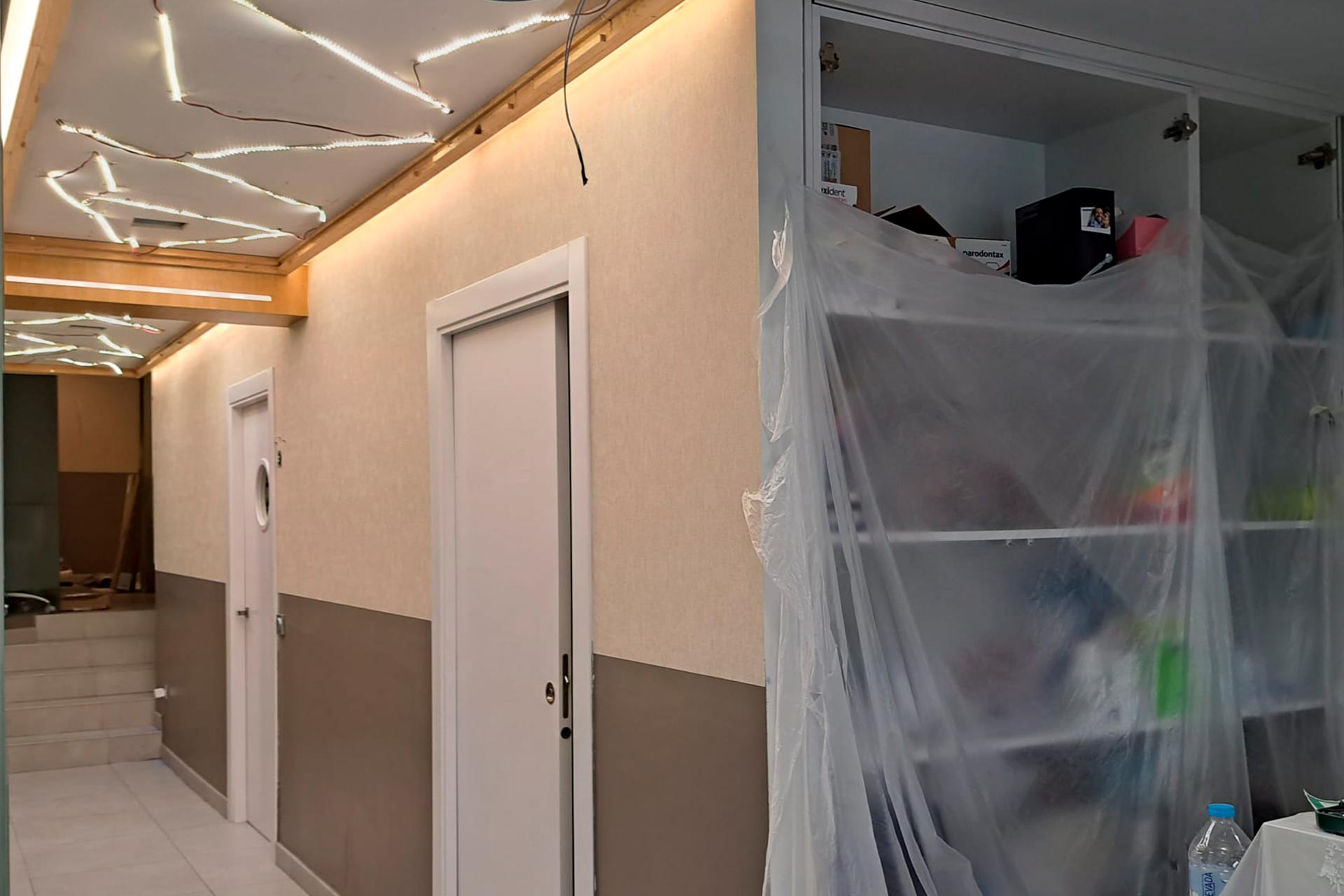 Un pasillo en renovación, con tiras de luz expuestas en el techo y una estantería cubierta de plástico a la derecha.