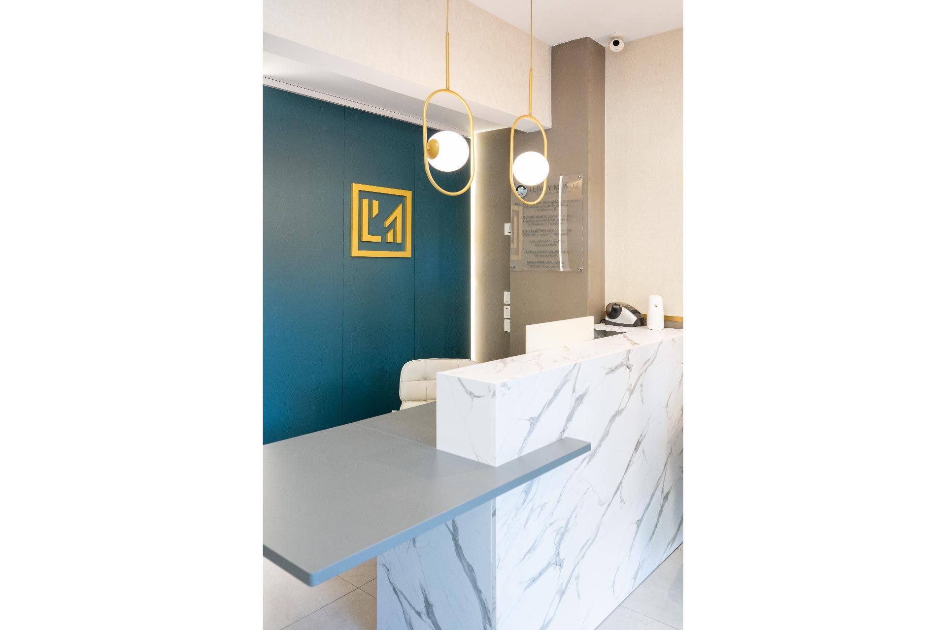 Área de recepción moderna con un escritorio de mármol, una pared con detalles en azul y un logotipo geométrico. Dos lámparas colgantes cuelgan del techo.