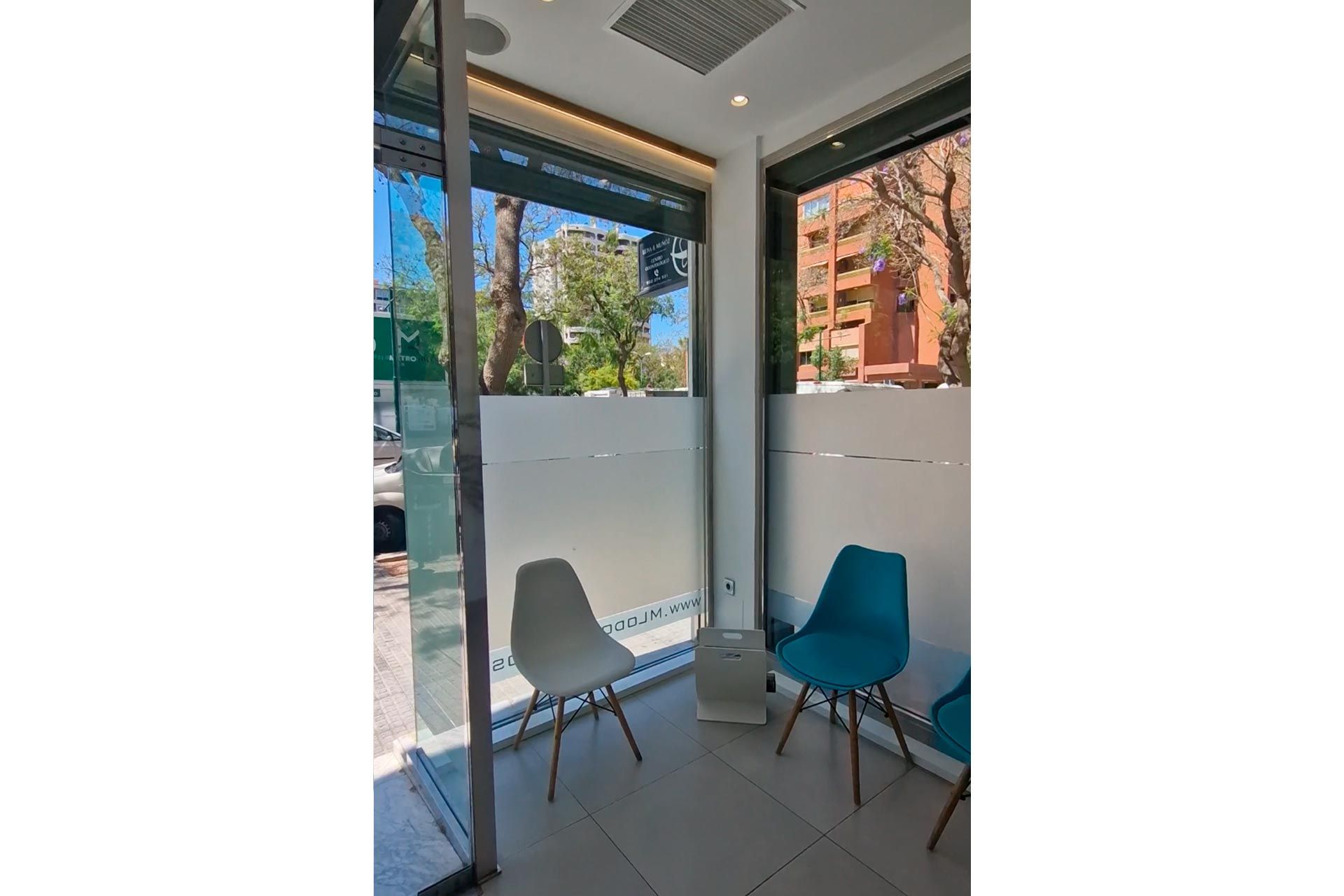 Dos sillas azules en un rincón de una habitación con paredes de vidrio con paneles esmerilados, con vista a una calle con árboles y edificios afuera.