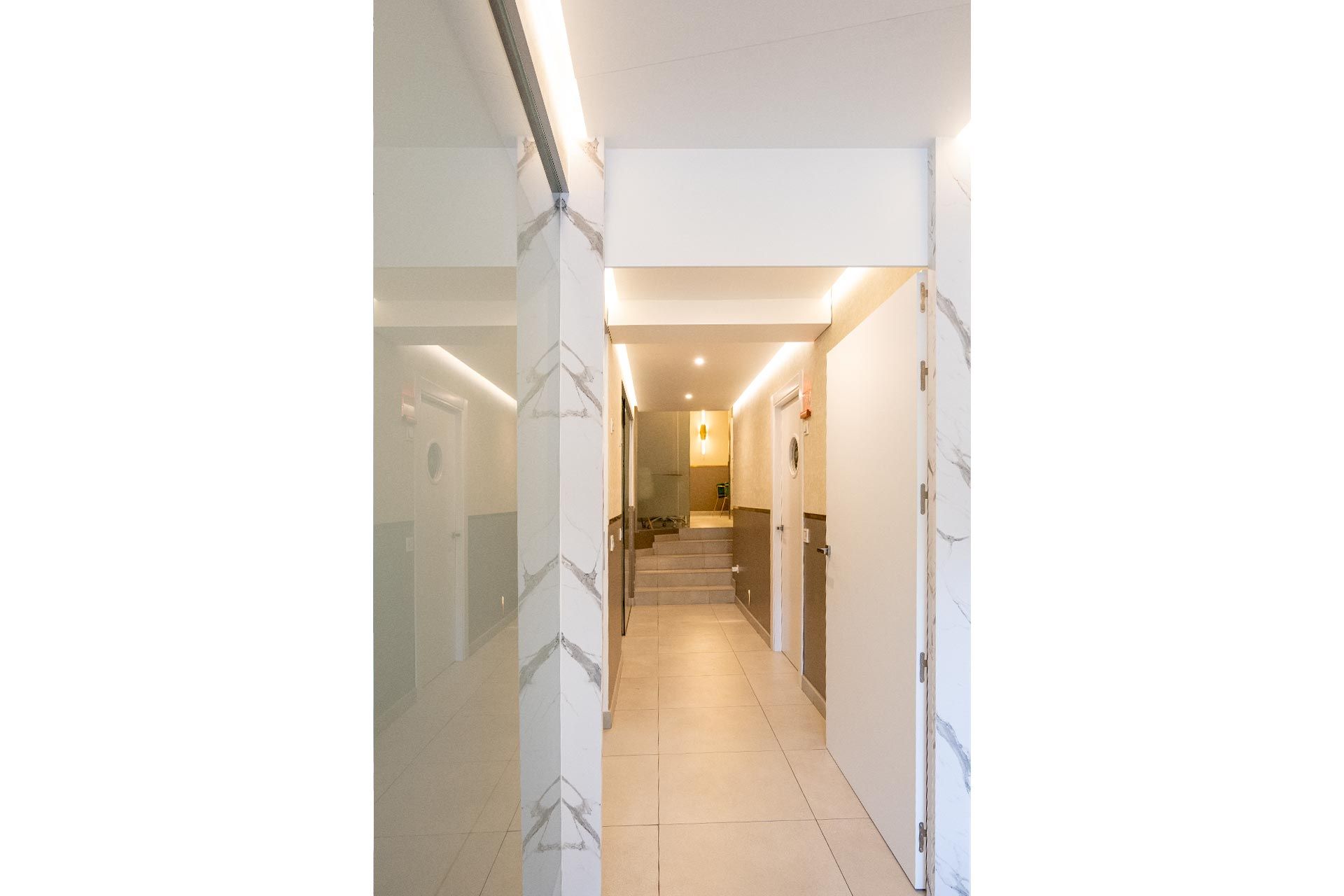 Pasillo luminoso con paredes y suelo de azulejos, que conduce a unas escaleras y a una zona cálidamente iluminada.