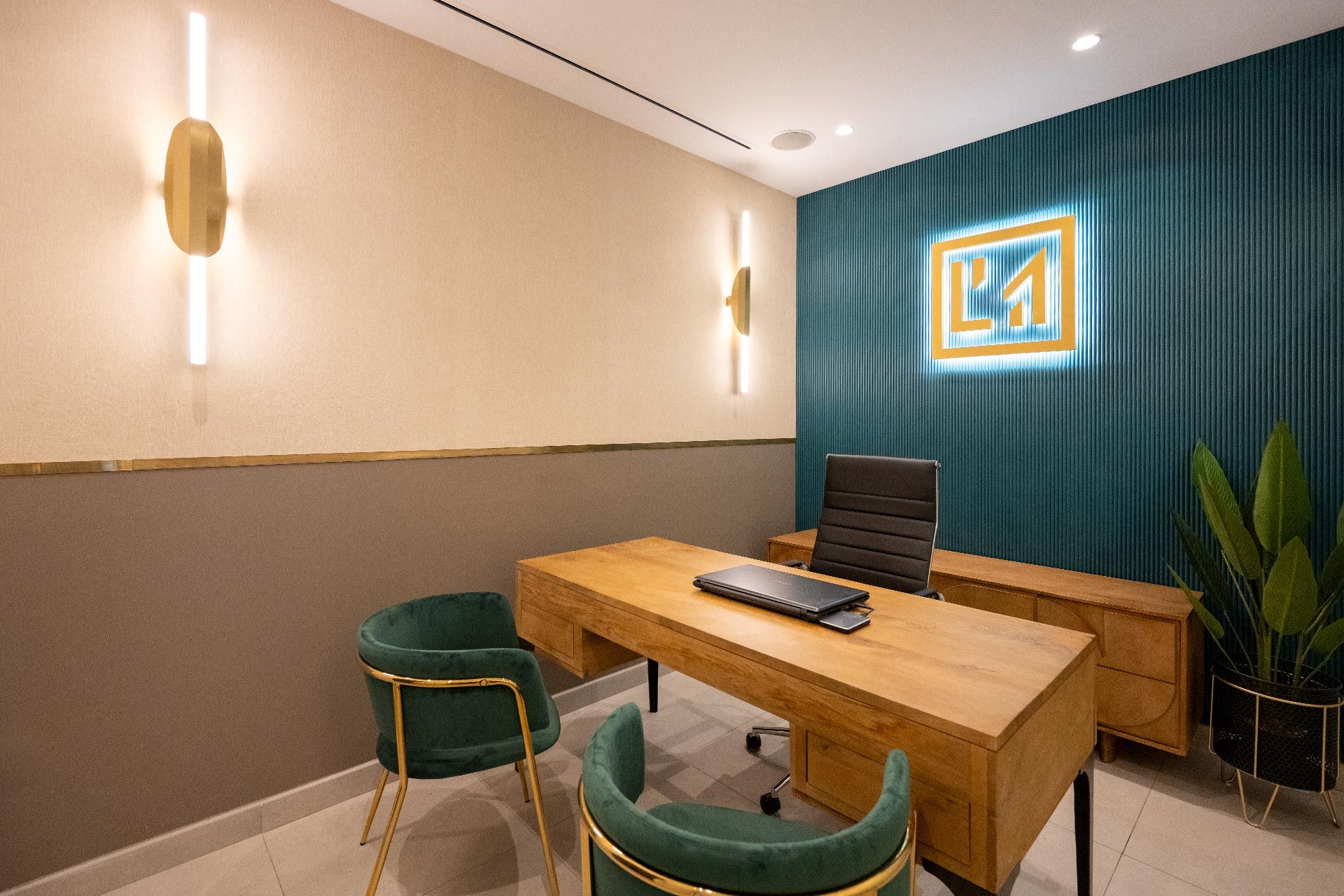 Oficina moderna con un escritorio de madera, una computadora portátil y una silla negra. Dos sillas verdes frente al escritorio. Las paredes tienen detalles dorados y un logotipo en la sección verde azulado. Hay dos apliques de pared y una planta en maceta.