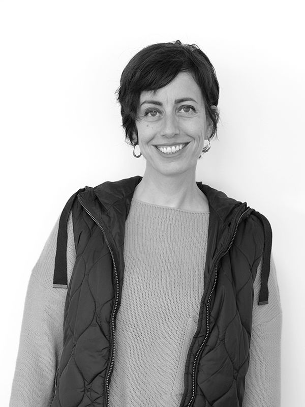 Blamnca-Cano Persona con cabello corto sonriendo, vistiendo un chaleco acolchado sobre un suéter, de pie sobre un fondo blanco liso.