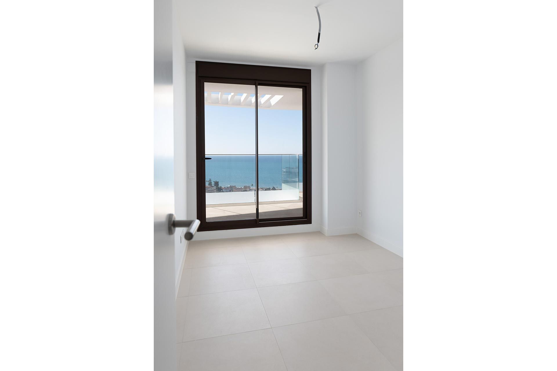 Habitación vacía minimalista con suelo de baldosas claras, una ventana con vistas al mar y una lámpara de techo inacabada.