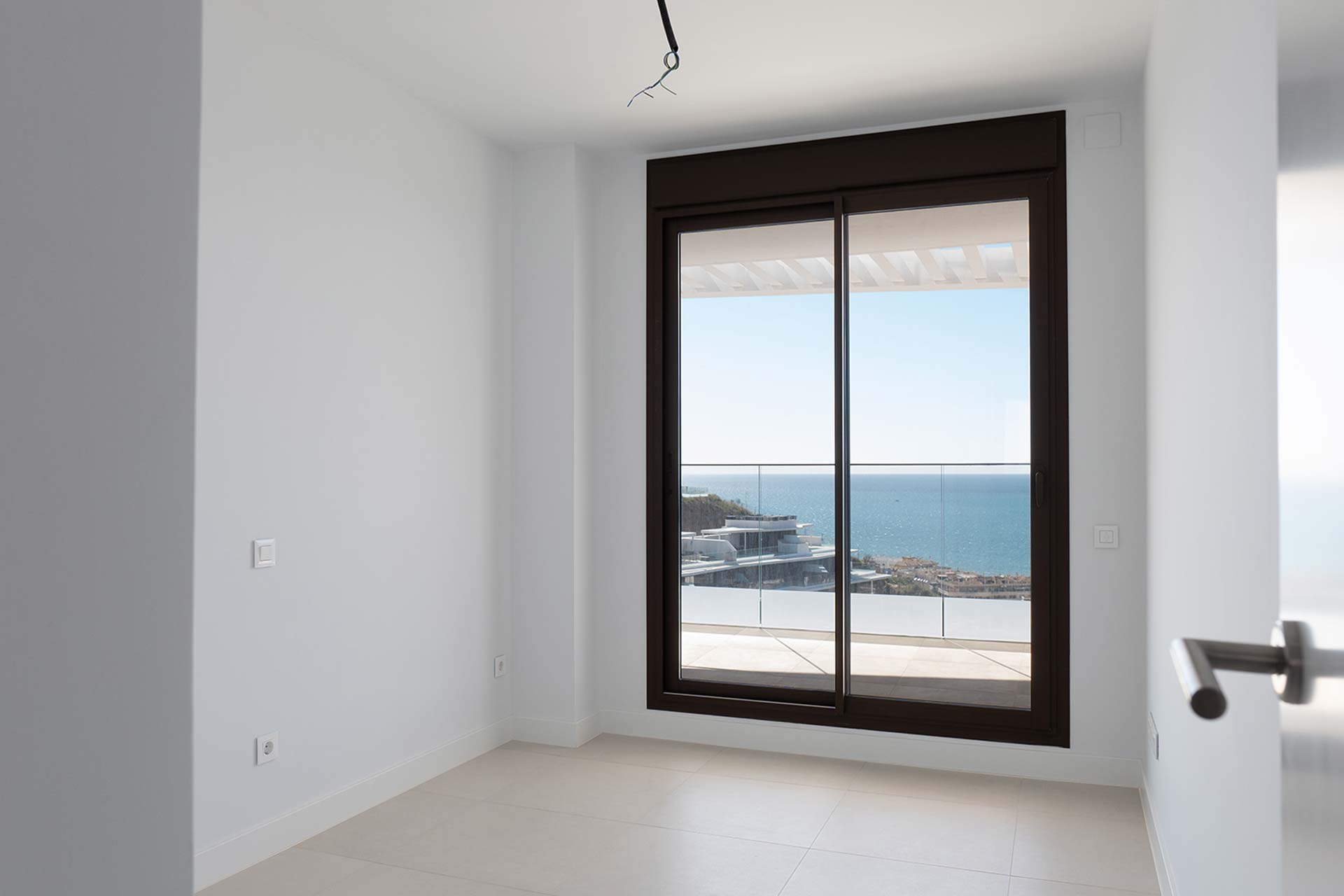 Habitación blanca y vacía con una gran puerta de cristal que da a un balcón con vistas al mar y a los edificios costeros bajo un cielo despejado.