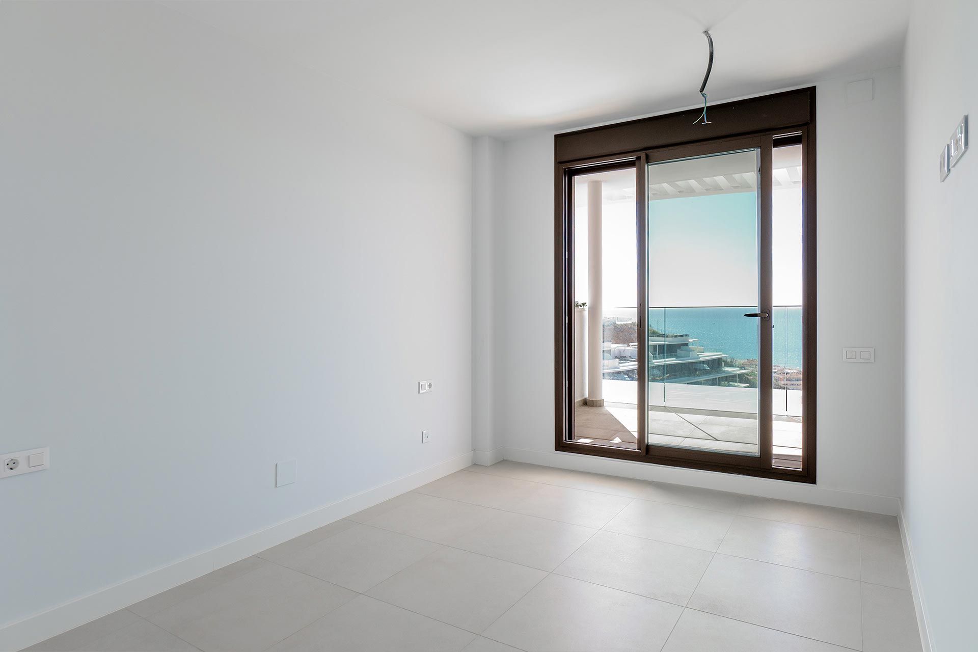 Habitación blanca vacía con suelo de baldosas, gran puerta de cristal y vista a un balcón con vistas al mar y a los edificios costeros bajo un cielo despejado.