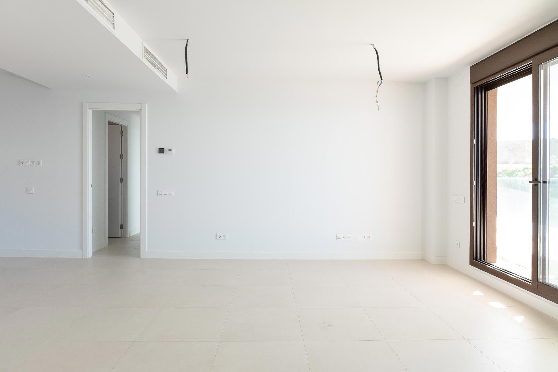 Habitación vacía, sin amueblar, con paredes blancas, suelo de baldosas grandes, una puerta abierta y puertas correderas de cristal que dejan pasar la luz natural. Los cables del techo están a la vista.