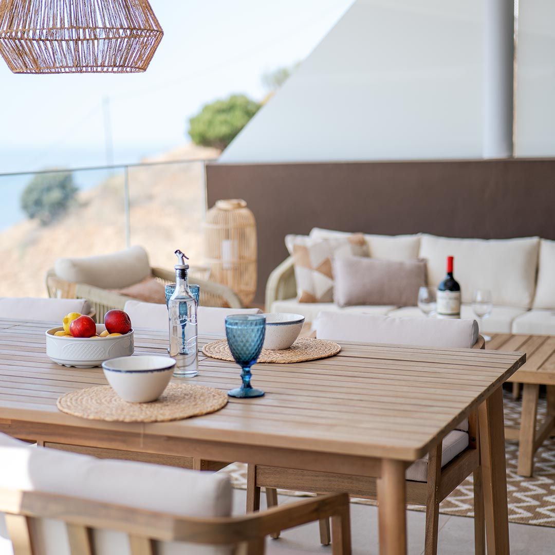 Un moderno patio exterior en Rincón de la Victoria cuenta con una mesa de comedor de madera con vajilla, cristalería y fruta, además de una acogedora zona de estar con un sofá y una botella de vino, perfecta para disfrutar de las vistas desde tu elegante Ático.