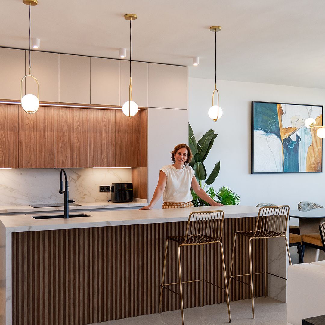 Interiorismo-Atico-Benalmadena-BlancaCano-Cocina-000 Una mujer se encuentra detrás de una moderna isla de cocina con paneles verticales de madera, encimeras de mármol y taburetes dorados; las lámparas colgantes y un gran cuadro abstracto completan la elegancia del espacio.