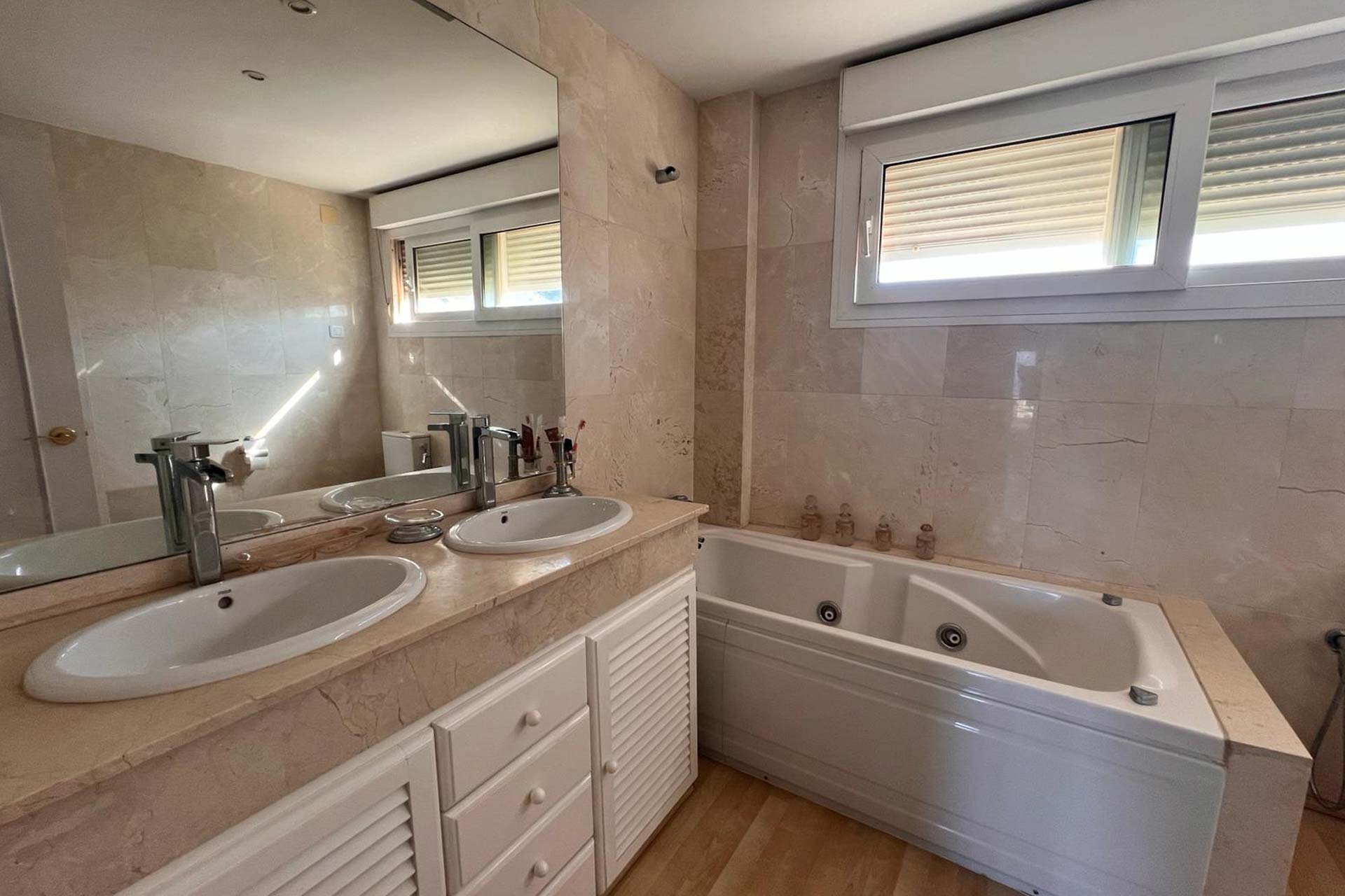 Cuarto de baño con doble lavabo, un gran espejo, un armario blanco, una bañera empotrada con chorros y una ventana con persianas que deja pasar la luz natural.