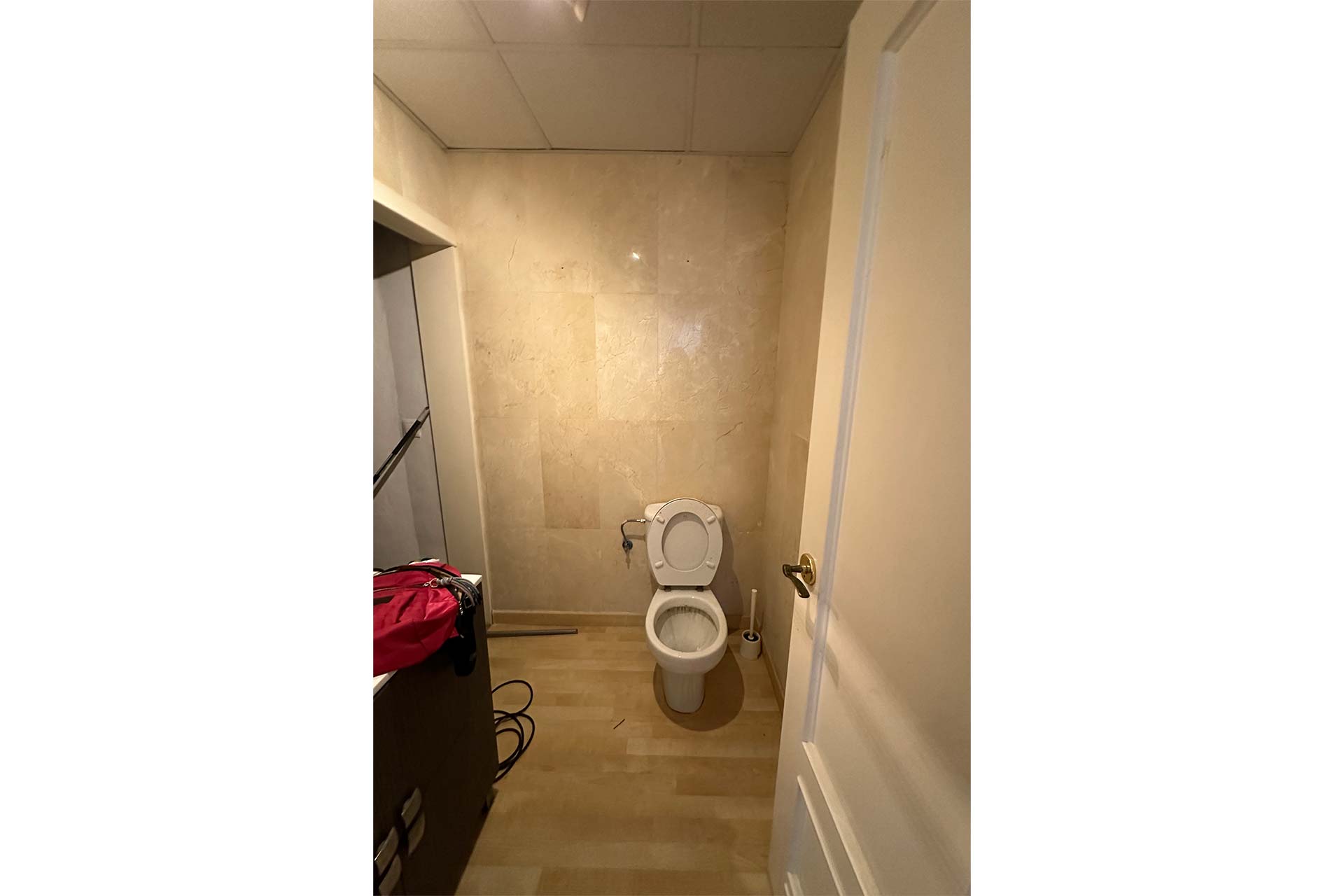 Un pequeño cuarto de baño con paredes y suelos de color beige, con un inodoro blanco, un armario a la izquierda y una puerta abierta en primer plano.
