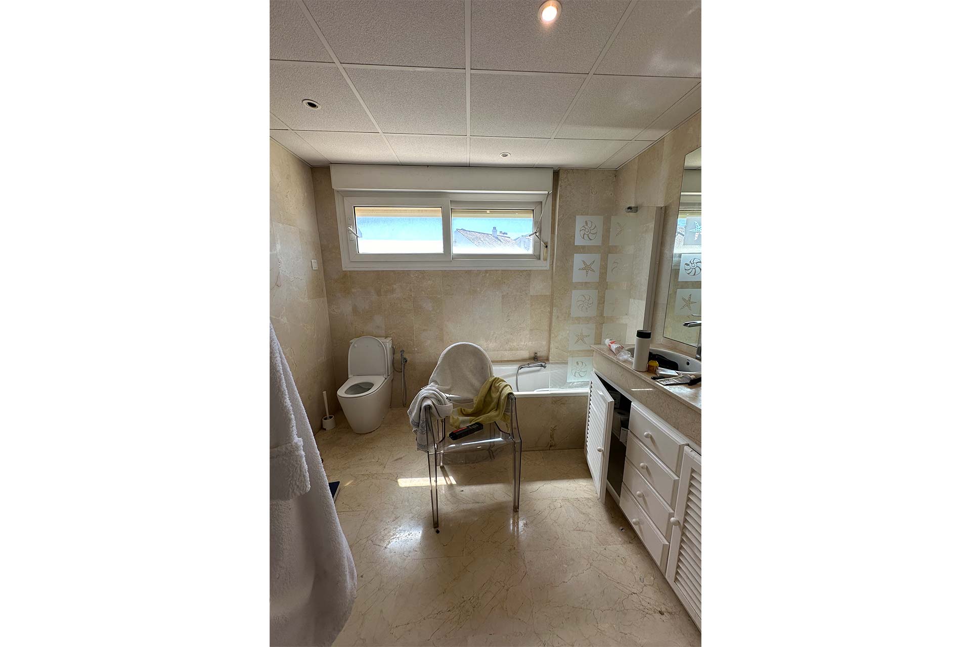 Un cuarto de baño con inodoro, bañera, espejo grande, lavabo y una silla con una toalla encima, bajo un techo con iluminación empotrada.