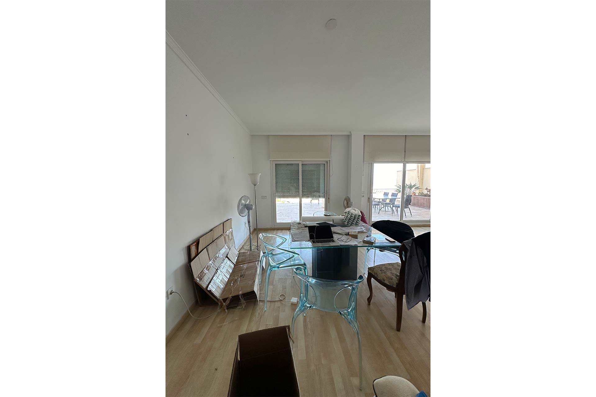 Un comedor con una mesa de cristal, sillas transparentes, un banco de madera, papeles esparcidos y vistas a un patio a través de grandes ventanales.