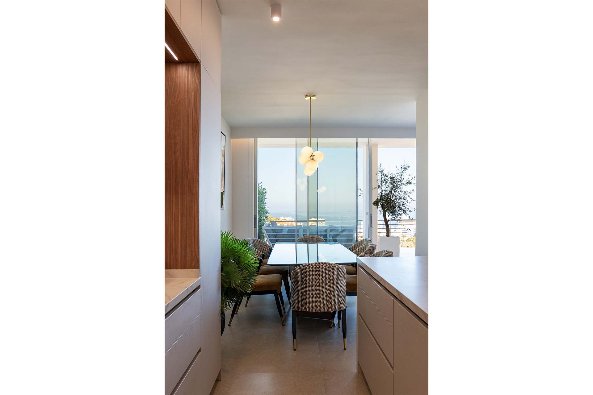 Moderno comedor con mesa de cristal, seis sillas, lámpara colgante, grandes ventanales y vistas a los árboles y al mar exterior.