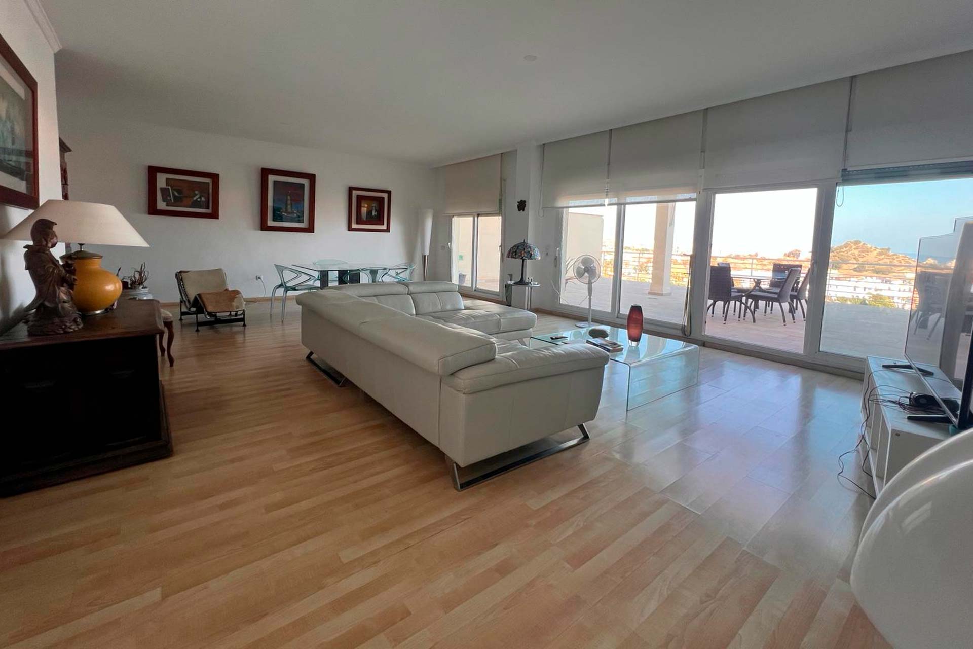 Amplio salón con sofá blanco, suelo de madera, obras de arte en la pared, mesa de comedor de cristal y grandes ventanales que dan a una terraza con asientos al aire libre y vistas panorámicas.