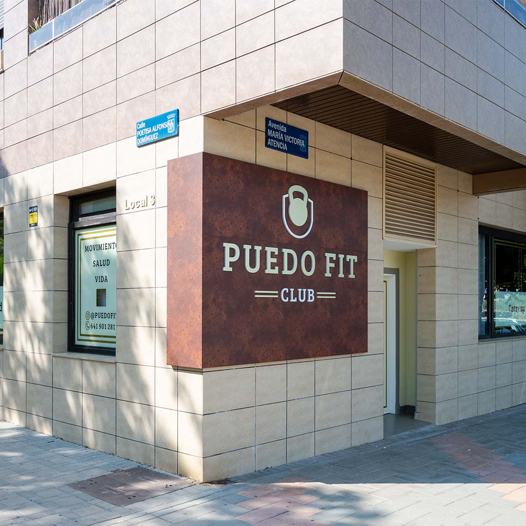 Vista exterior de un edificio con un gran cartel en el que se lee "Puedo Fit Club", señales de tráfico de la Avenida María Victoria Atencia y de la Plaza Alonso Vergara, y un borrador automático visible junto a la entrada.
