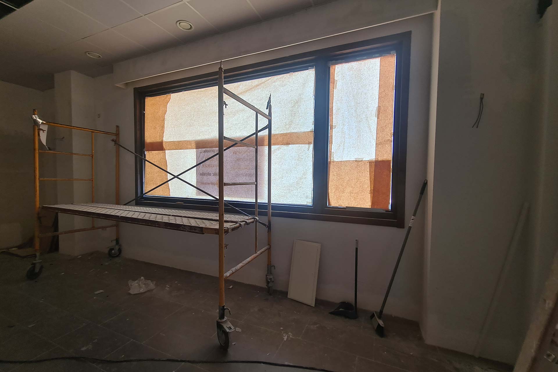 En una habitación inacabada, con materiales de construcción y herramientas en el suelo, se colocan andamios frente a una gran ventana cubierta con papel de estraza.