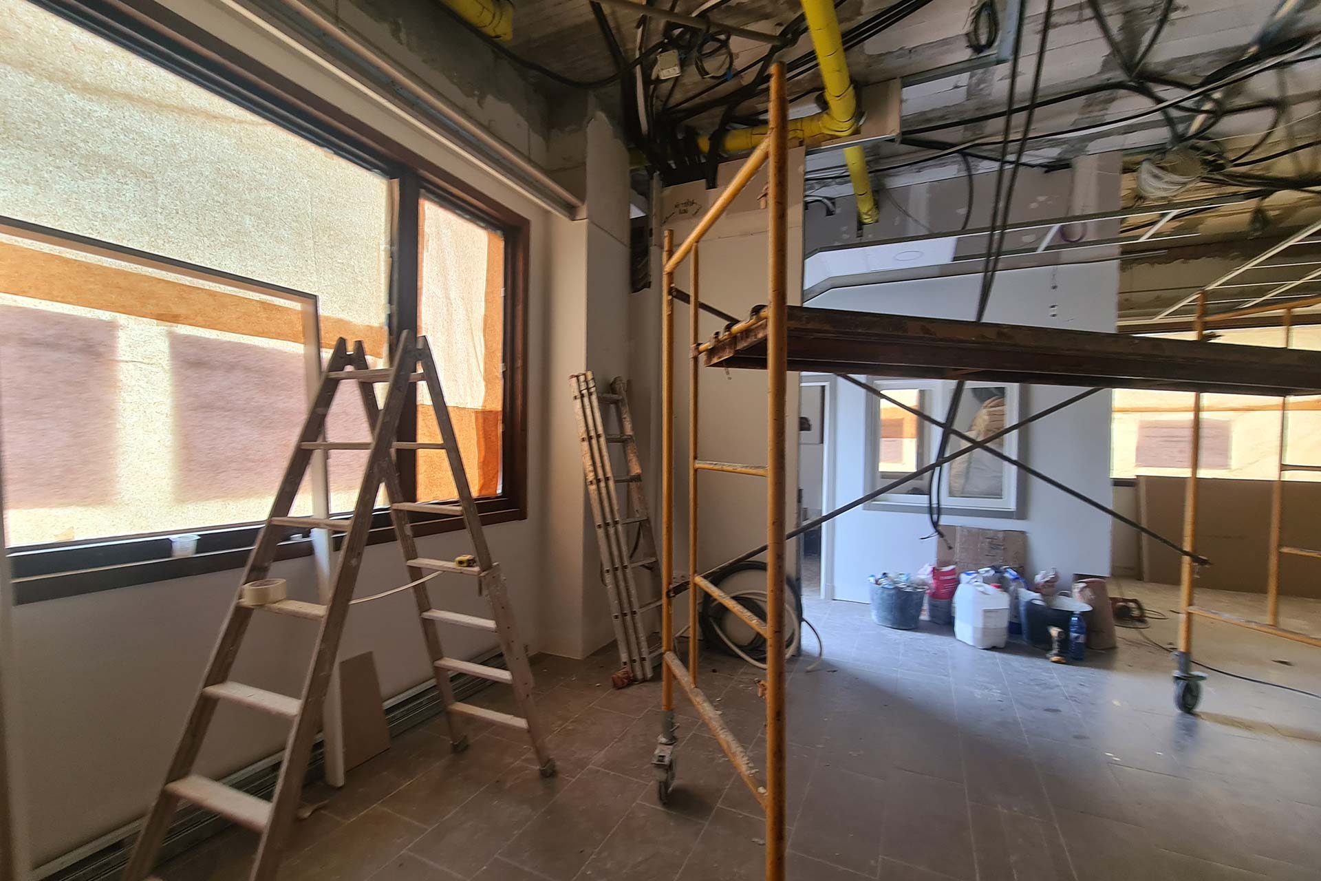 Habitación en renovación con escaleras, andamios, tuberías expuestas y materiales de construcción esparcidos por el suelo cerca de ventanas cubiertas con papel marrón.