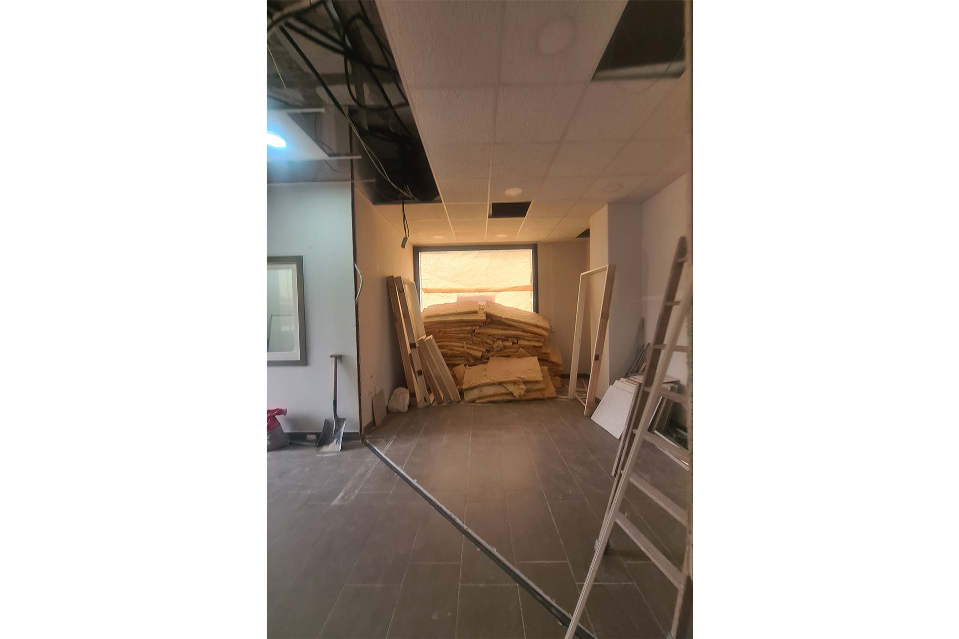 Una zona de construcción con escaleras, herramientas y una gran pila de paneles aislantes apilados cerca de una ventana. En el techo faltan baldosas y el cableado está al descubierto.