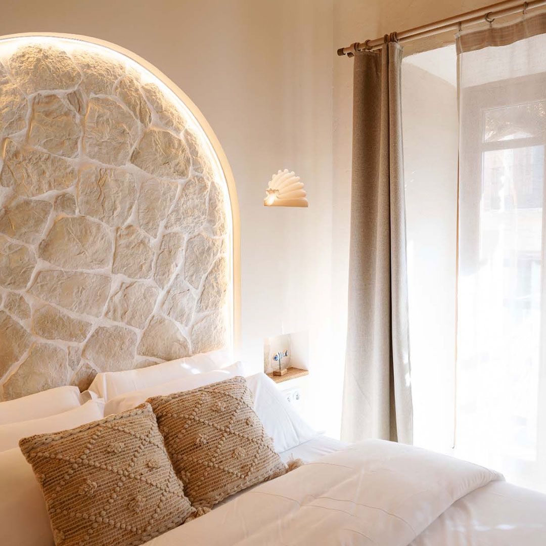 Un dormitorio con una cama grande, cortinas azules, luces colgantes de color naranja, una manta estampada, un cuadro colgado en la pared y una pared con detalles de azulejos.