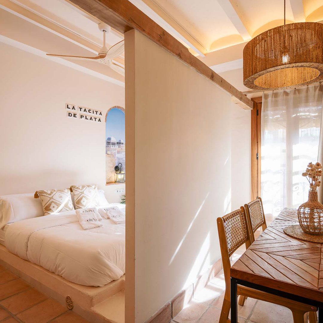 Un dormitorio con una cama grande, cortinas azules, luces colgantes de color naranja, una manta estampada, un cuadro colgado en la pared y una pared con detalles de azulejos.