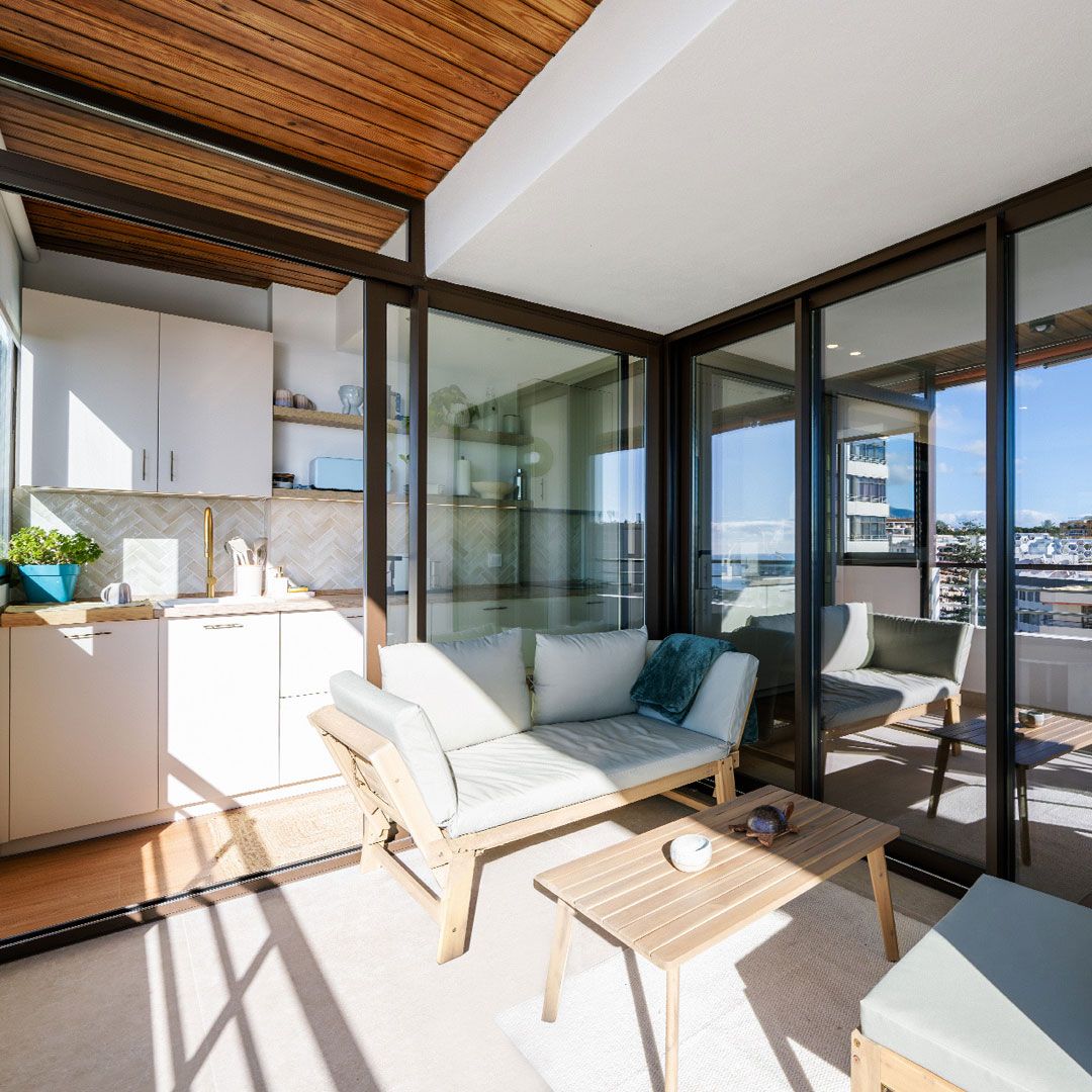 Moderno balcón con paredes de cristal, un sofá de madera acolchado, una mesa pequeña y una cocina exterior compacta con armarios y un fregadero, con vistas a un soleado paisaje urbano.