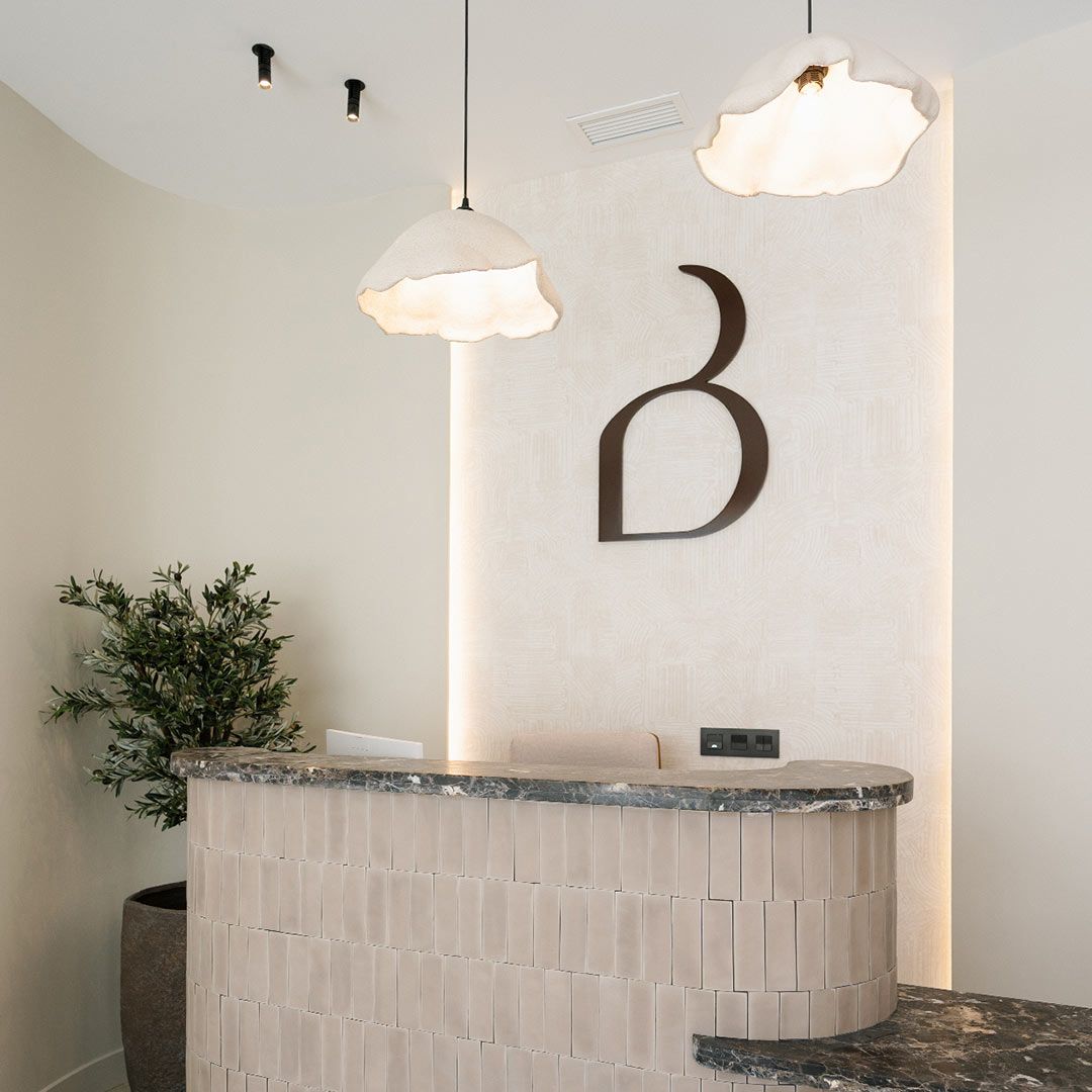 Un mostrador de recepción con azulejos curvados, una maceta, dos lámparas colgantes y un logotipo en la pared con una "b" estilizada crean un interior minimalista y moderno para la Clínica Burgos.