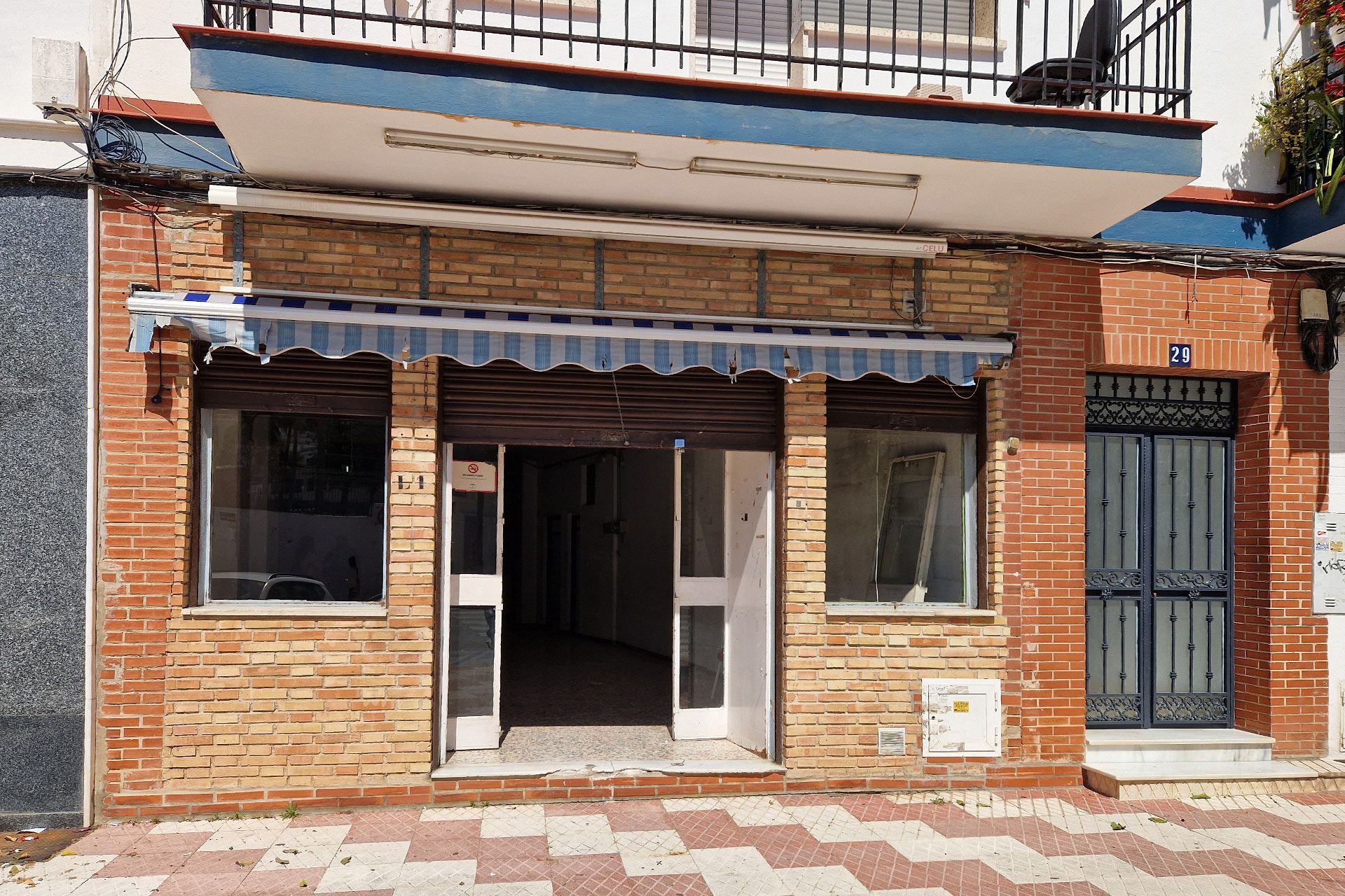Fachada cerrada con un toldo a rayas azules y blancas, dos ventanas frontales y una entrada abierta; pasillo de baldosas delante y una puerta vecina a la derecha.