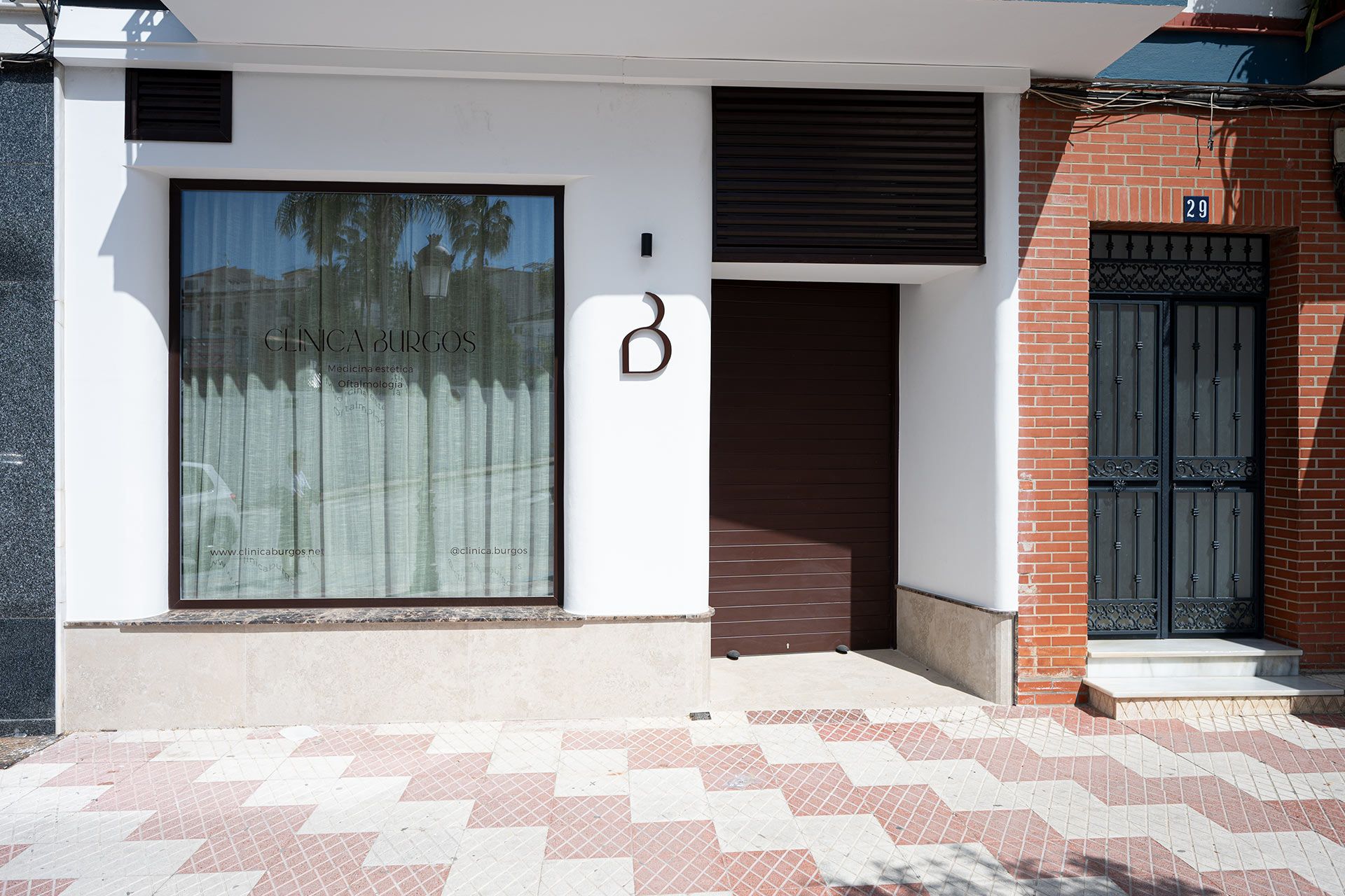 Vista de la calle de un moderno escaparate blanco con una gran ventana que muestra "CINCA BURGOS", una puerta marrón cerrada y una puerta de entrada de metal negro a la derecha marcada con el número 29.