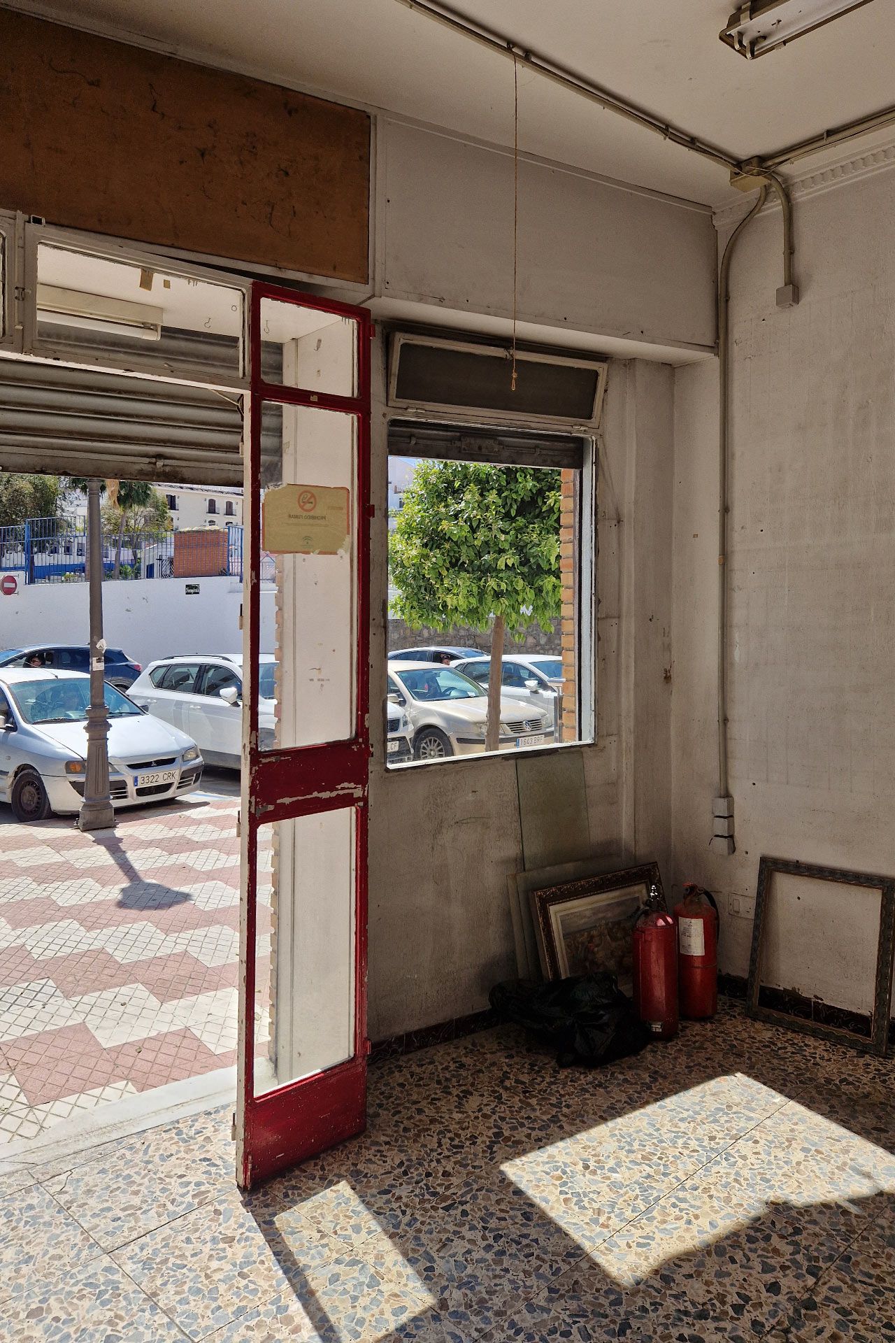 Una habitación vacía con una puerta de cristal de marco rojo abierta al exterior, varios coches aparcados a la vista, extintores y cuadros enmarcados apoyados en la pared.