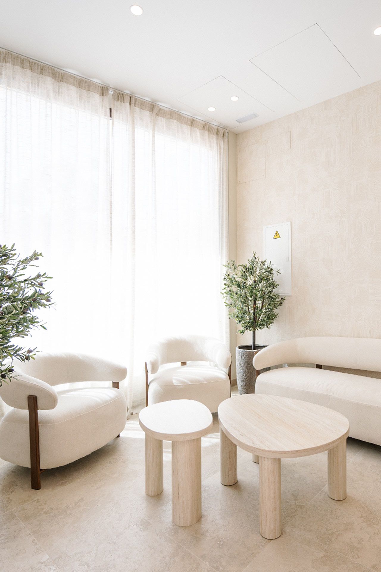 Salón minimalista con sillones color crema, un pequeño sofá, dos mesas de centro redondas, plantas en macetas y grandes cortinas transparentes que dejan pasar la luz natural.