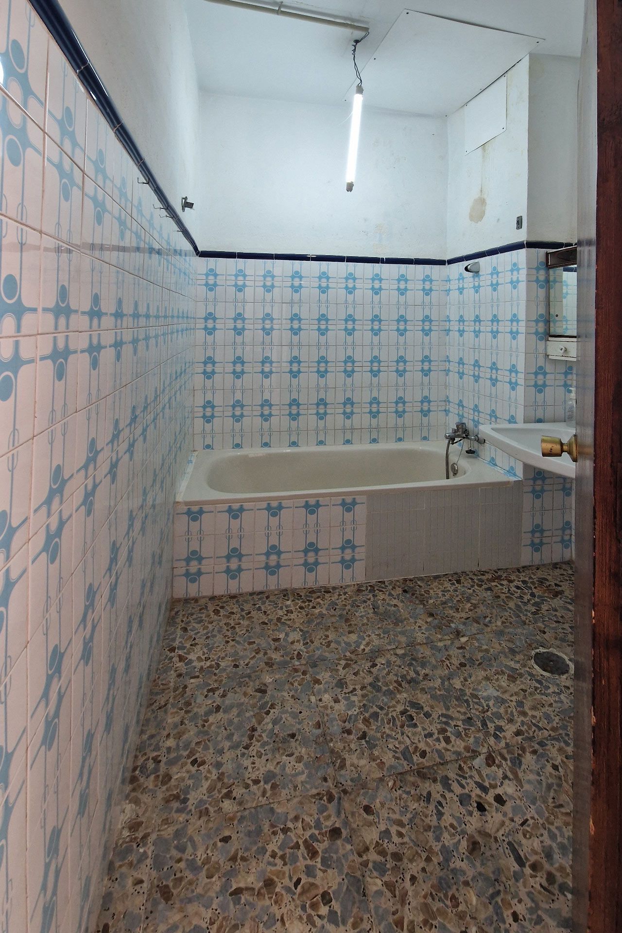 Un cuarto de baño con bañera, azulejos azules y blancos, suelo de piedra, lavabo y luz fluorescente.