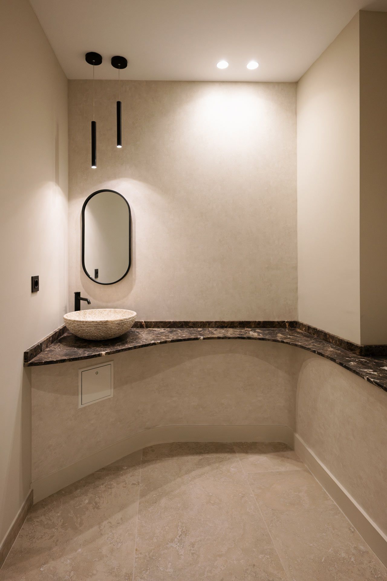 Moderno cuarto de baño con encimera de mármol oscuro, espejo ovalado, lavabo de piedra, lámparas colgantes y paredes y suelo de baldosas beige.