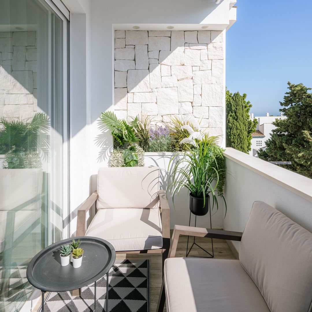 Balcón moderno con sillas acolchadas de color beige, una mesita negra con plantas en macetas, una alfombra geométrica y una pared de piedra blanca, con vistas a la vegetación y una vista parcial de los edificios a lo lejos.